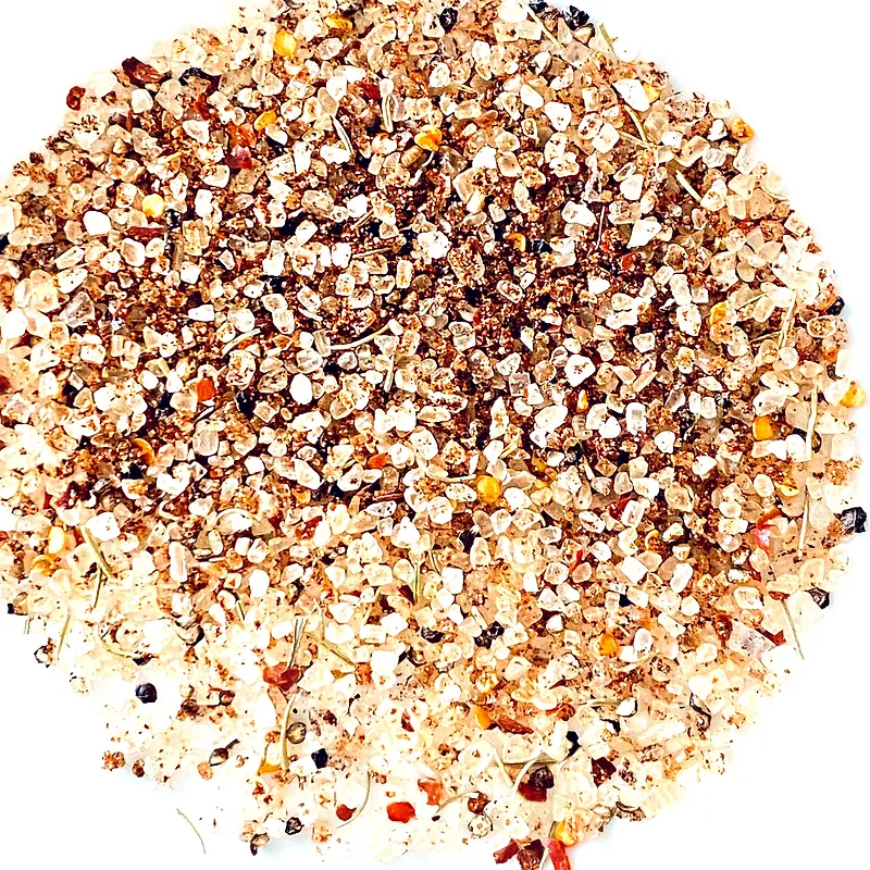 A close-up, overhead view of a coarse spice blend composed of small, irregular granules and flecks of herbs, all against a stark white background. The spices are a mix of light and dark brown, white, and some reddish-orange fragments, suggesting ingredients like salt, pepper, dried herbs, and possibly chili flakes or paprika.