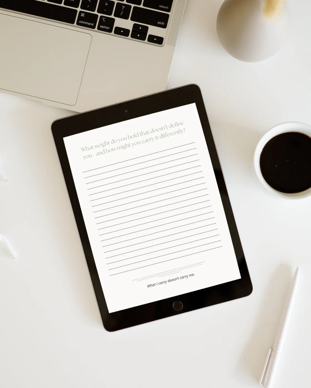 A tablet screen displaying a journal entry template with lined paper and a motivational quote is placed next to a laptop, a cup of coffee, and a pen on a white desk. The journal page asks "What weight do you hold that doesn't define you - and how might you carry it differently?" and concludes with "What I carry doesn't carry me."