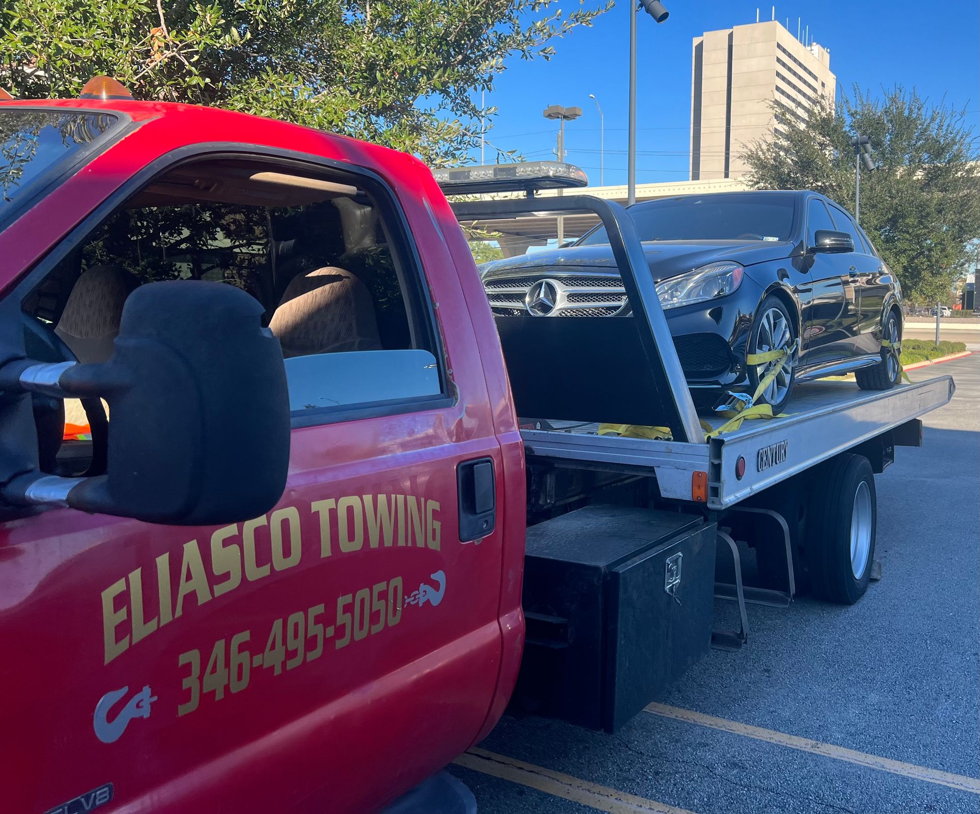 Eliasco Towing red flatbed truck securely transports a black Mercedes-Benz sedan in a commercial parking area, showcasing professional vehicle towing, recovery, and safe transportation services during daylight.