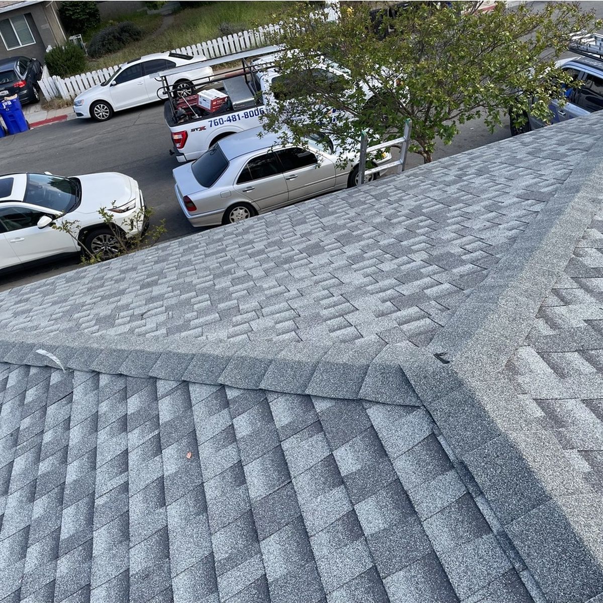 Close-up view of fresh gray asphalt shingles and hip cap detailing on a roof, looking down toward parked cars and work trucks on a residential street.