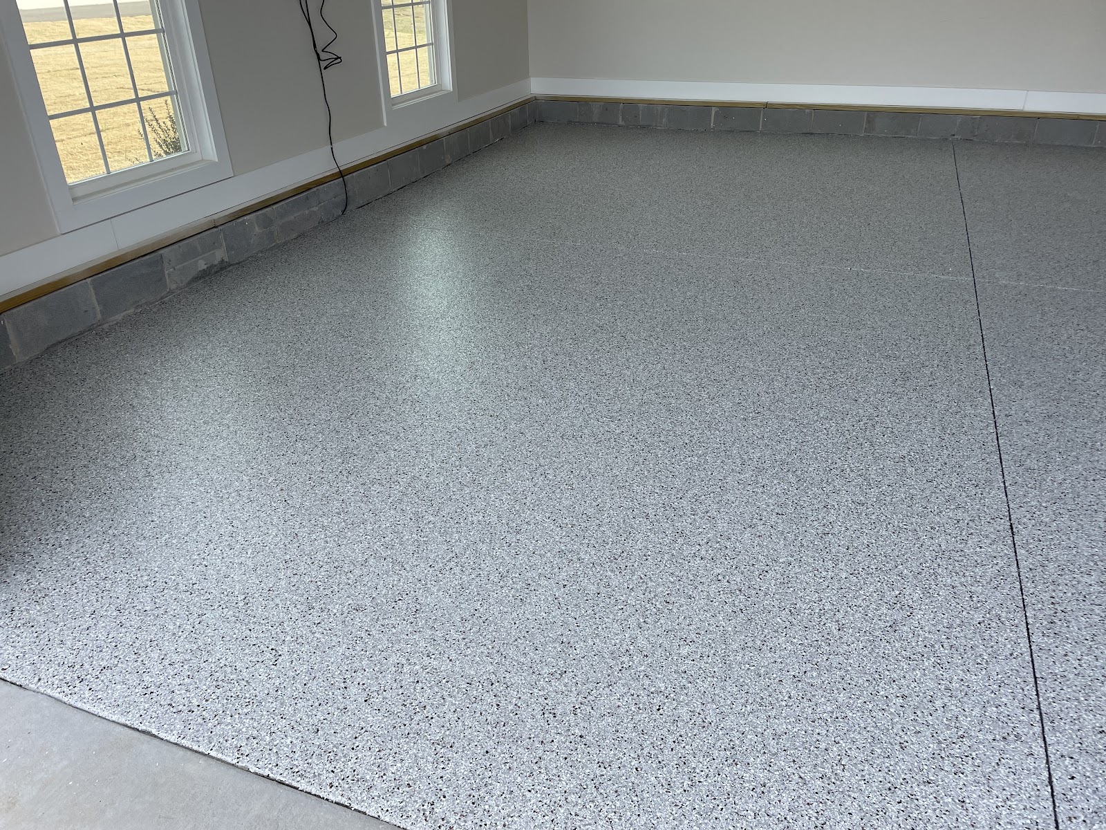 A speckled grey epoxy floor with a smooth, seamless finish is shown in a room with two windows and concrete block walls at the base. The floor has a faint reflection from the overhead lighting and a single seam running vertically down the right side of the image. The windows offer a view of dry, yellowed grass and a clear sky.