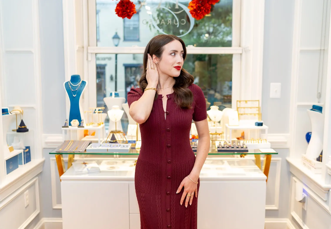Burgundy button-front dress is styled with delicate jewelry inside Eliana Curated. Bright displays showcase necklaces, rings, and bracelets at this boutique in Old Town Alexandria, Virginia. Visit us.