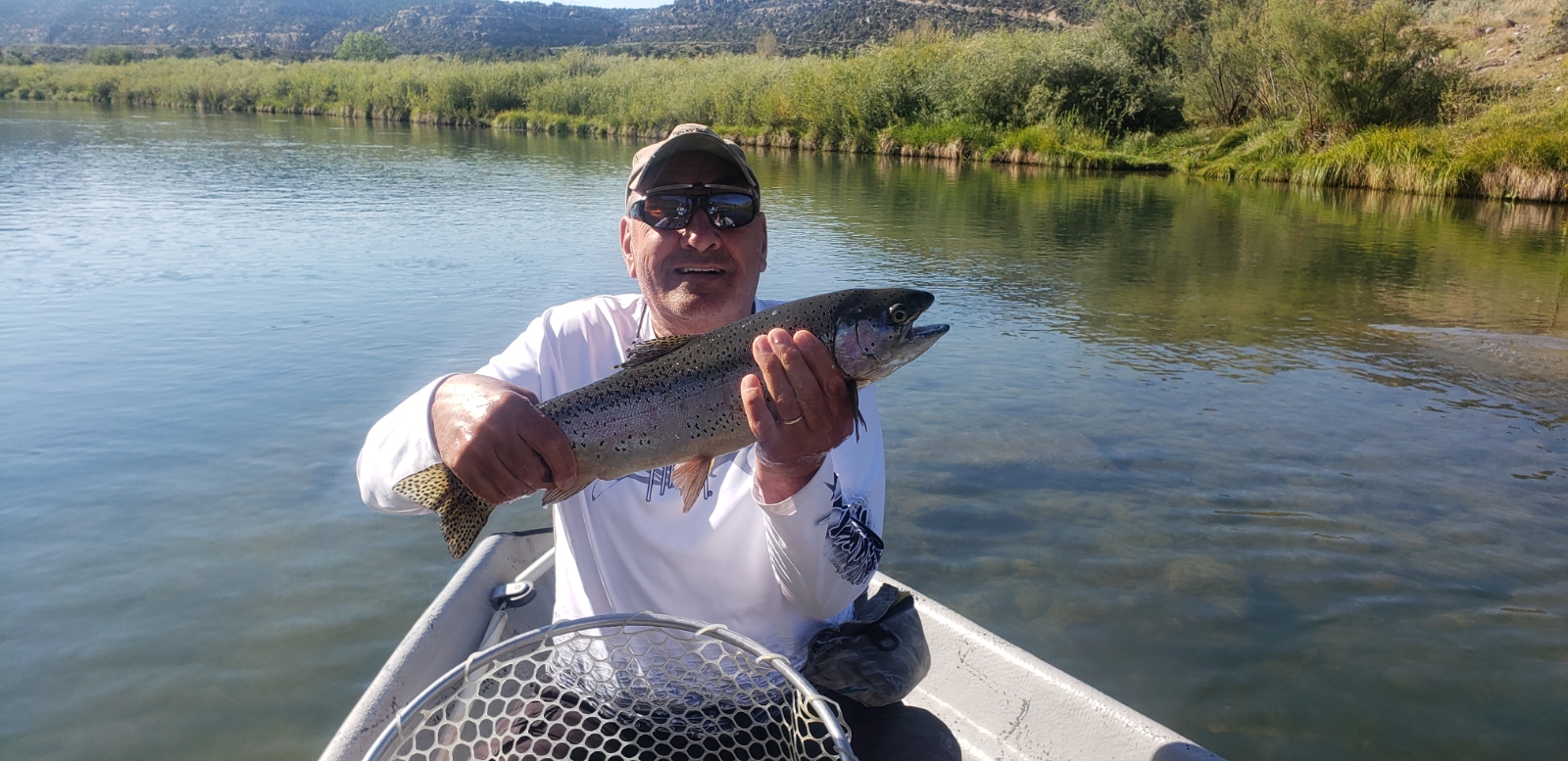 A man in a boat holds up a large rainbow trout he just caught, smiling at the camera. He is wearing a long-sleeved white shirt, sunglasses, and a baseball cap. The boat is on a calm river with trees and hills in the background. A fishing net is visible in the foreground.