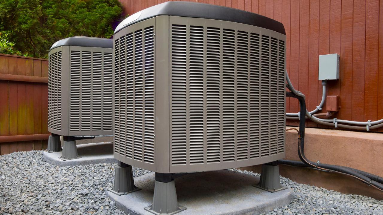 An outdoor air conditioning condenser unit is pictured on a concrete slab surrounded by grass. The unit is a large metal box with vents and a fan on top, and a series of fins on the sides. The fins are coated with a thin layer of dust and debris. The unit is connected to a set of pipes and wires that run along the ground towards the building. The sky above is blue with scattered clouds