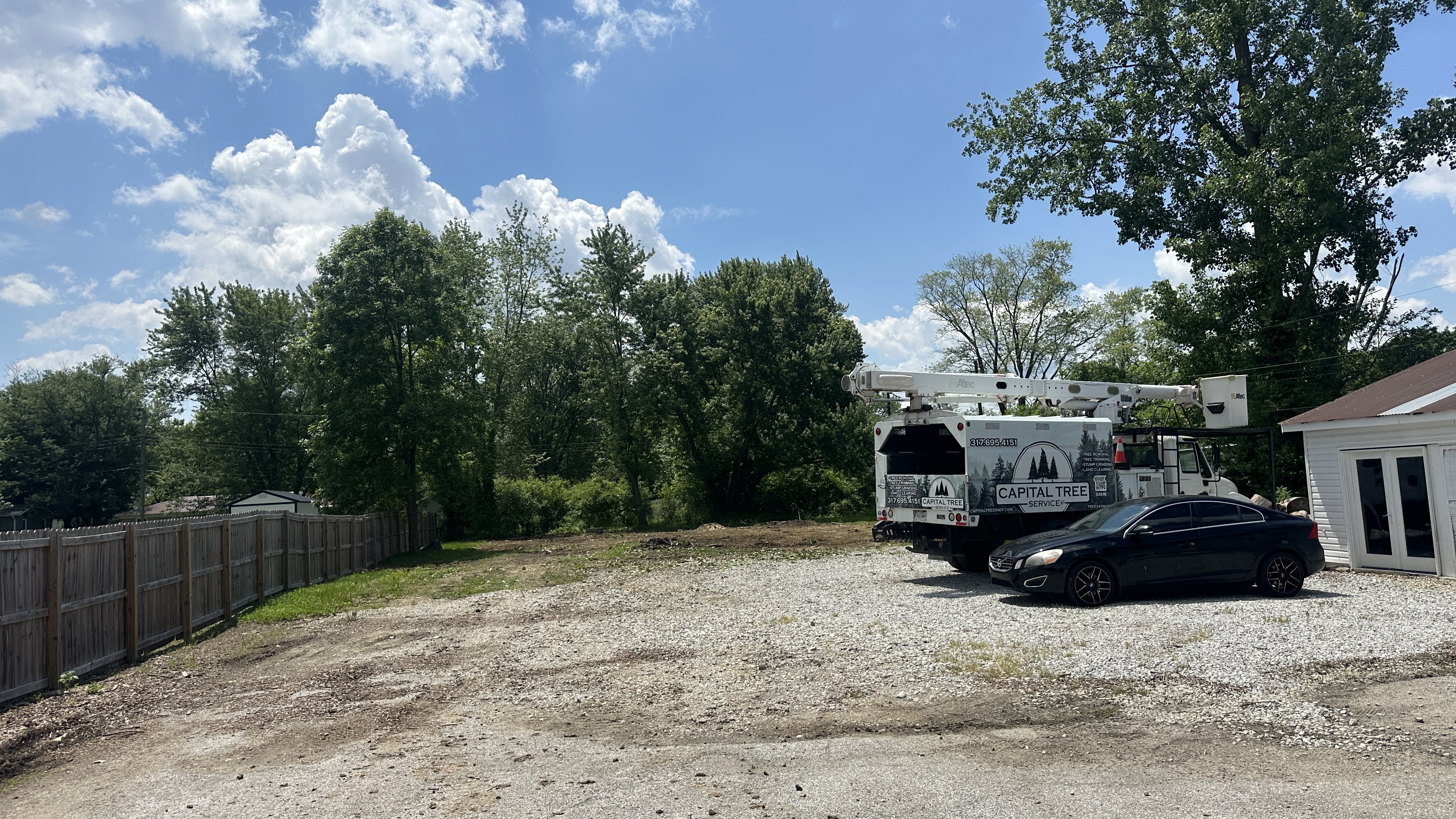 A Capital Tree Service truck and a black car are parked on a gravel lot next to a wooden fence and a white building under a bright blue sky with fluffy white clouds. The truck has a bucket lift and is branded with the company's name and phone number. Trees and foliage are visible in the background.
