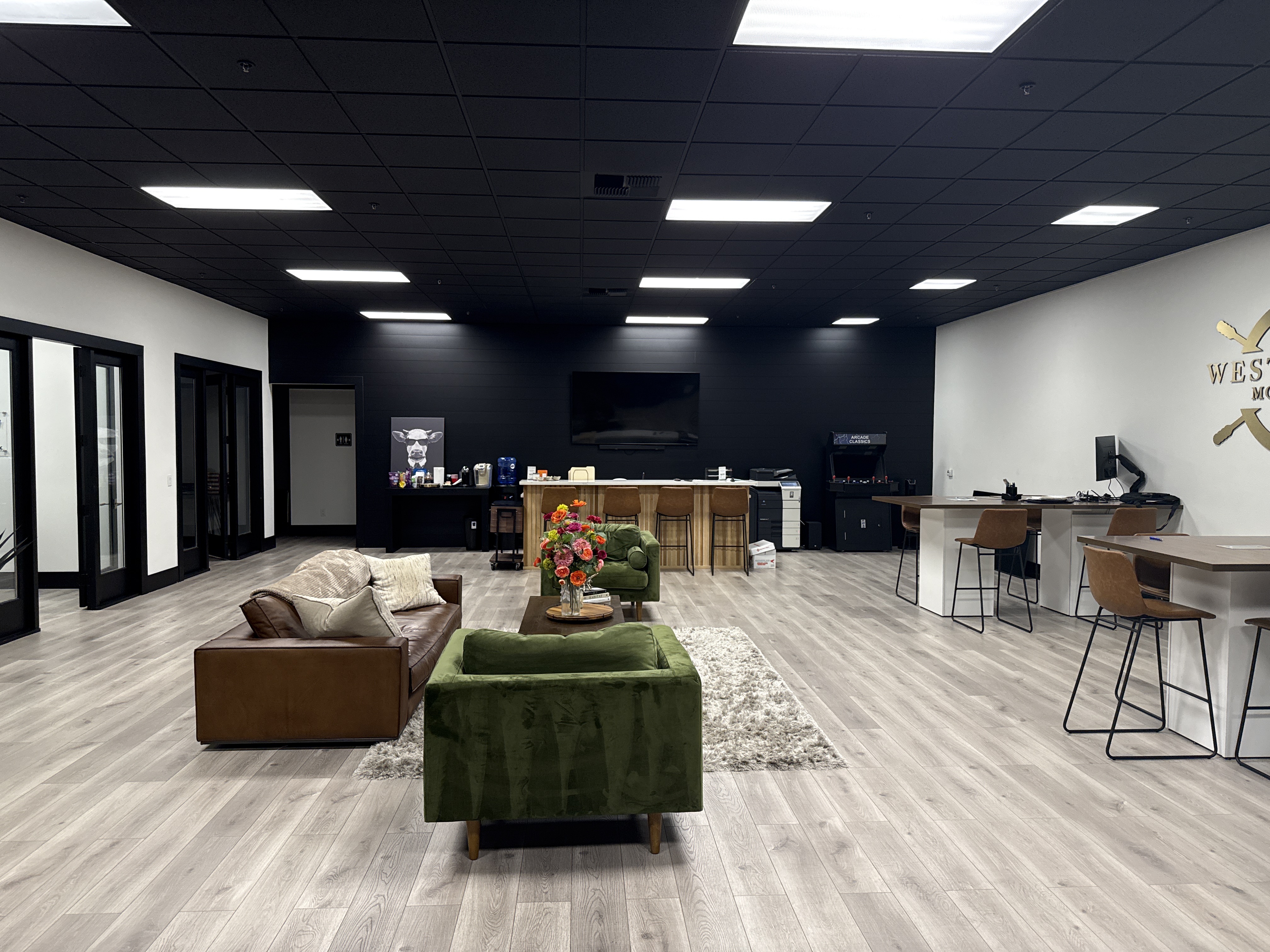 A spacious, modern office reception area featuring a lounge area with plush seating, a high-top bar with stools, desks with computers, and a distinctive arcade cabinet. The walls are painted black and white, and the ceiling is black with recessed lighting.