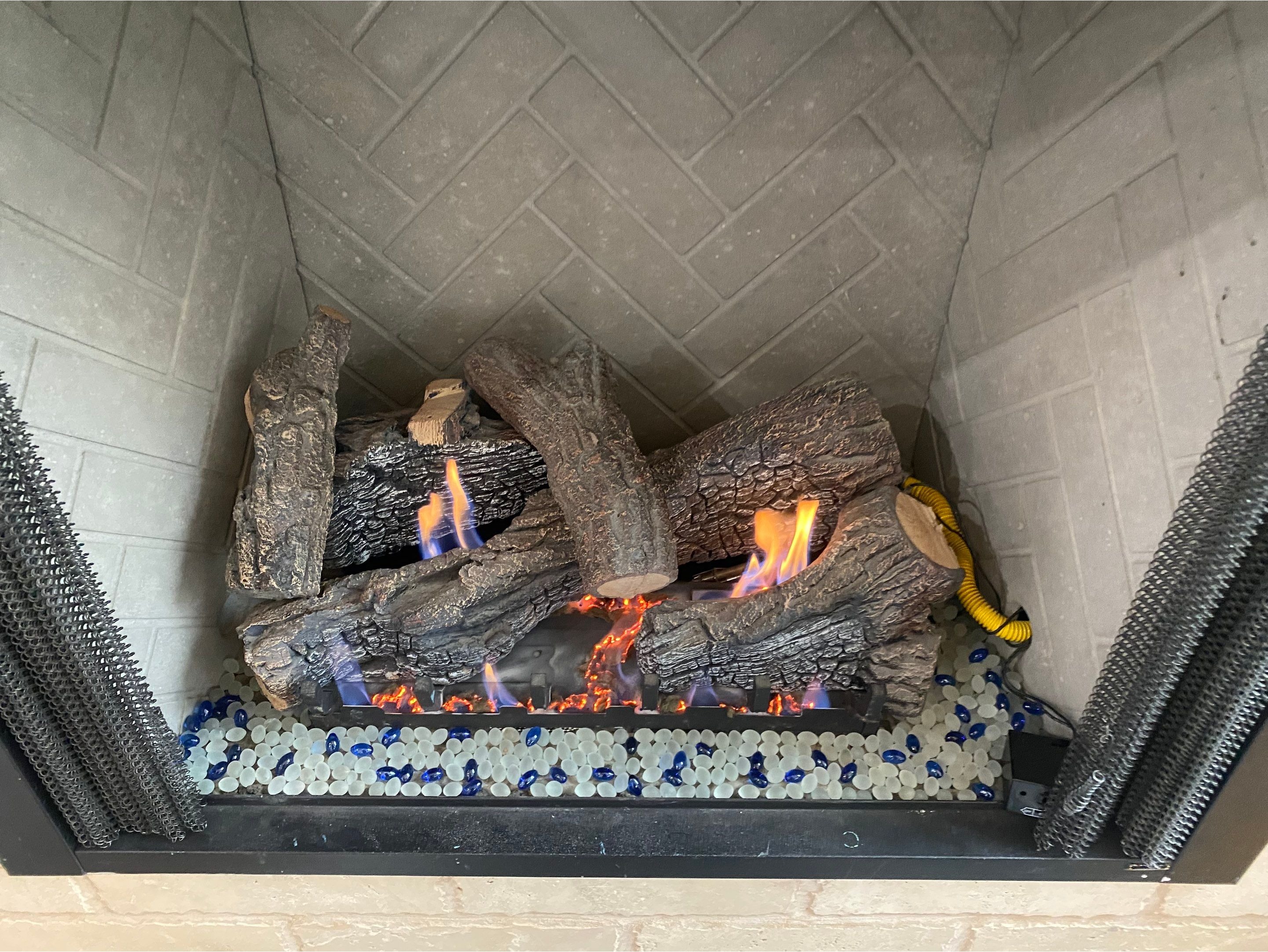 Gas log set burning over a bed of white and blue decorative fire glass, set against a herringbone-pa 