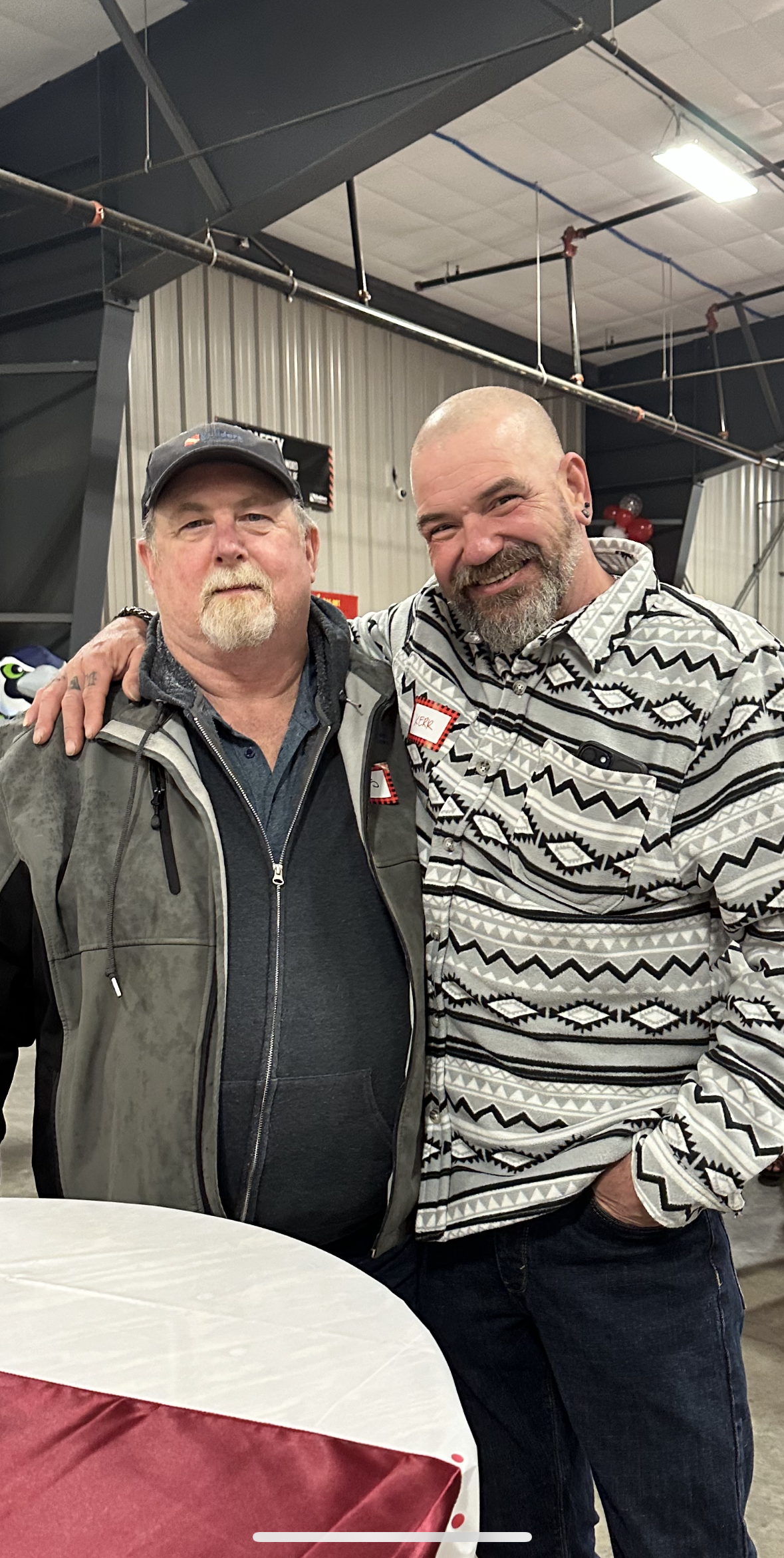 Two men are standing side by side, smiling and posing for the camera at an indoor event. The man on the left is wearing a dark jacket and a baseball cap, while the man on the right has his arm around the other’s shoulder and is dressed in a patterned button-up shirt. Both appear relaxed and happy, with name tags visible on their chests. The background shows an industrial interior with metal walls and exposed pipes, suggesting the event is taking place in a warehouse or manufacturing facility. A table with a red and white tablecloth is partially visible in the foreground.