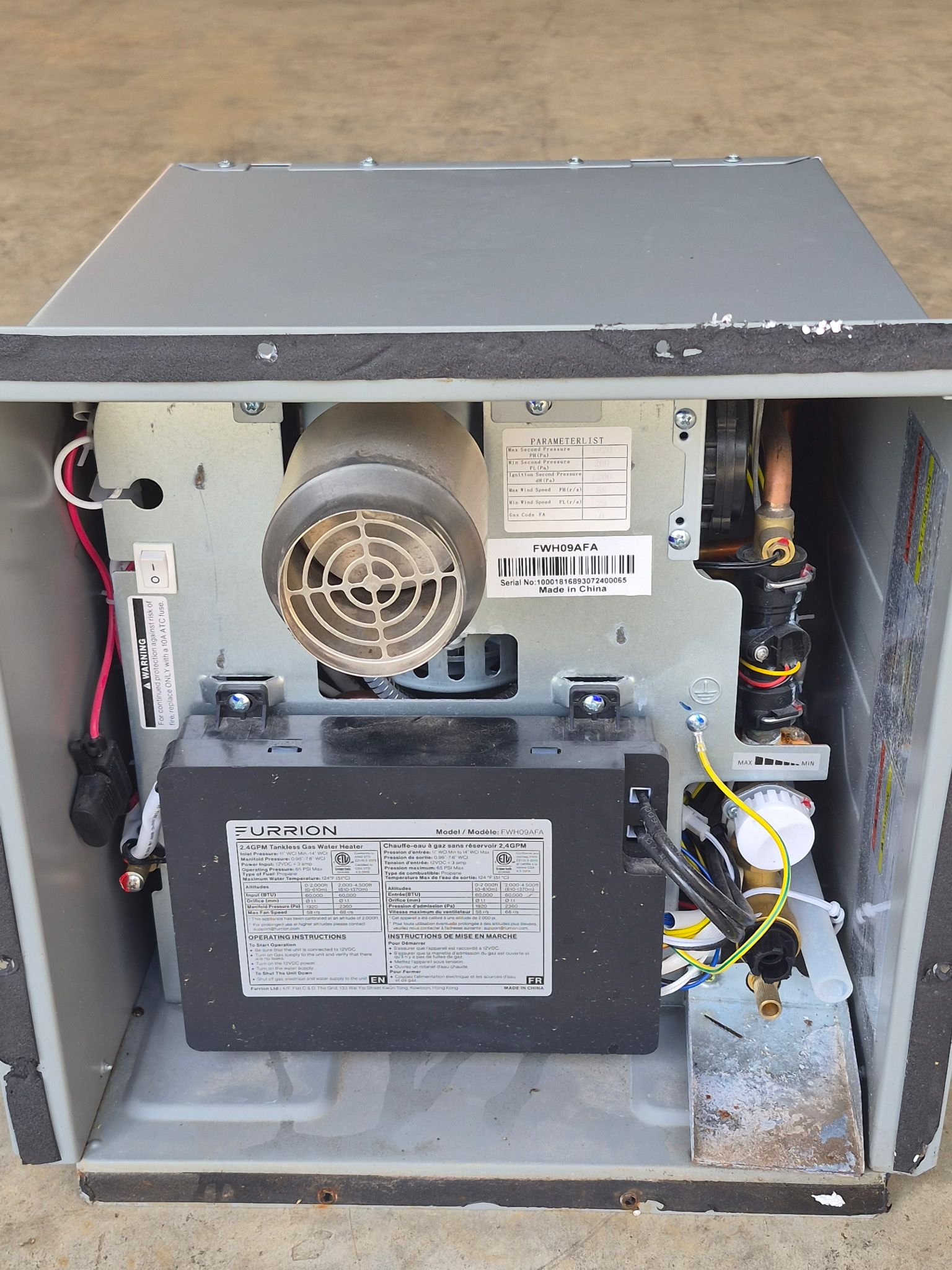 Open Furrion tankless RV water heater housing reveals blower assembly, wiring, copper piping, valves, control components, and labeled service panel, showing internal equipment during inspection, troubleshooting, or repair work nearby.