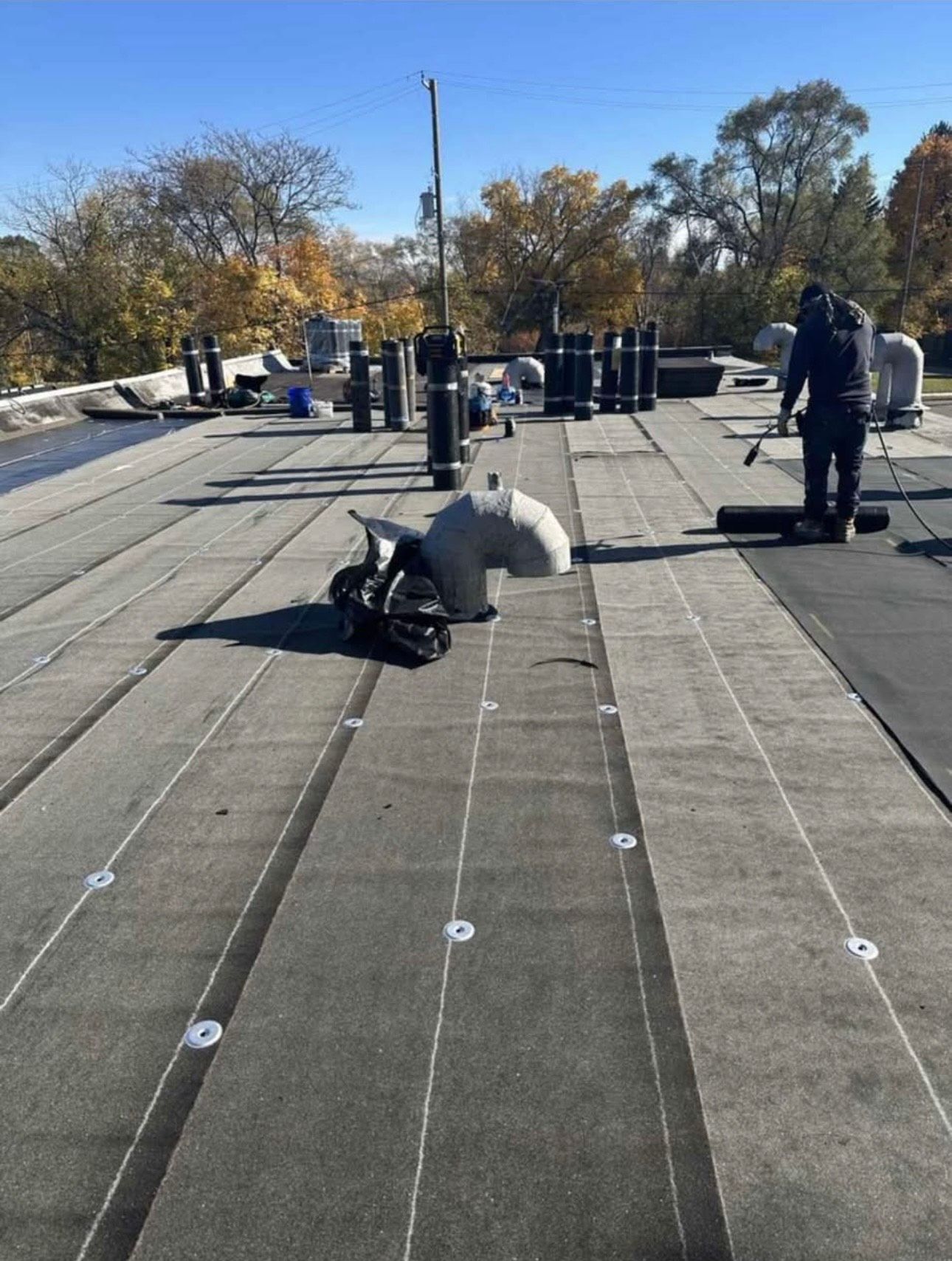 A worker performs torch-down application on a flat roof crowded with numerous vent pipes. Sections of dark membrane are installed, showing fastening discs. Tools rest nearby under sunny skies.
