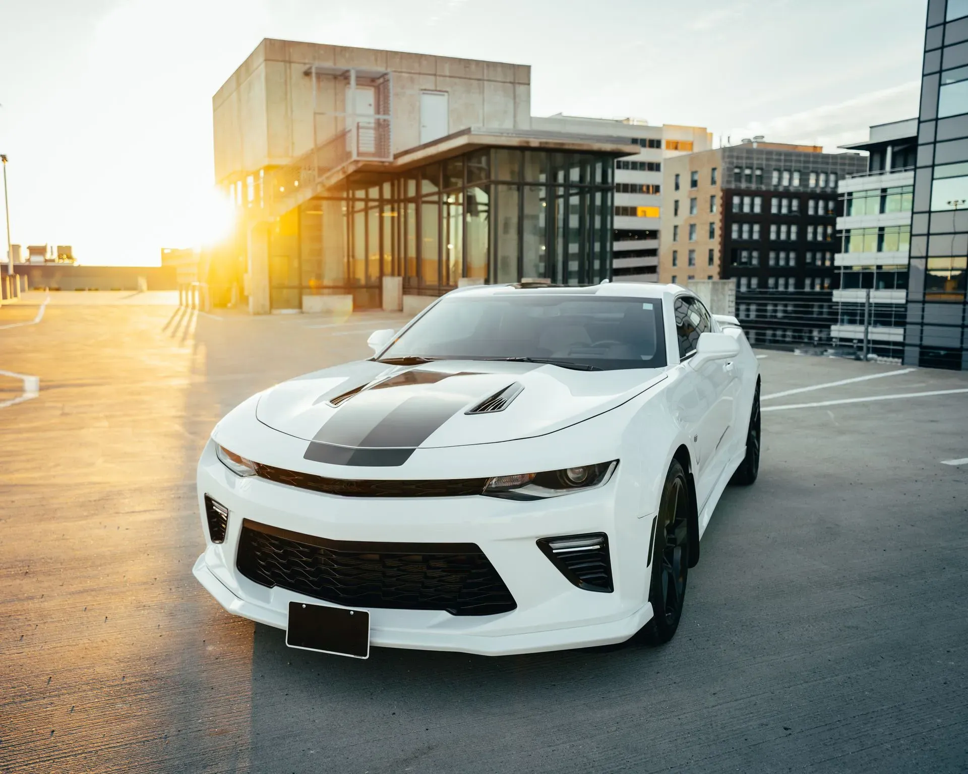 A white Chevrolet Camaro parked on a concrete surface, possibly a rooftop parking lot, with modern buildings in the background during sunset. The car features black racing stripes on the hood. The sun is setting to the left, casting a warm glow on the scene.