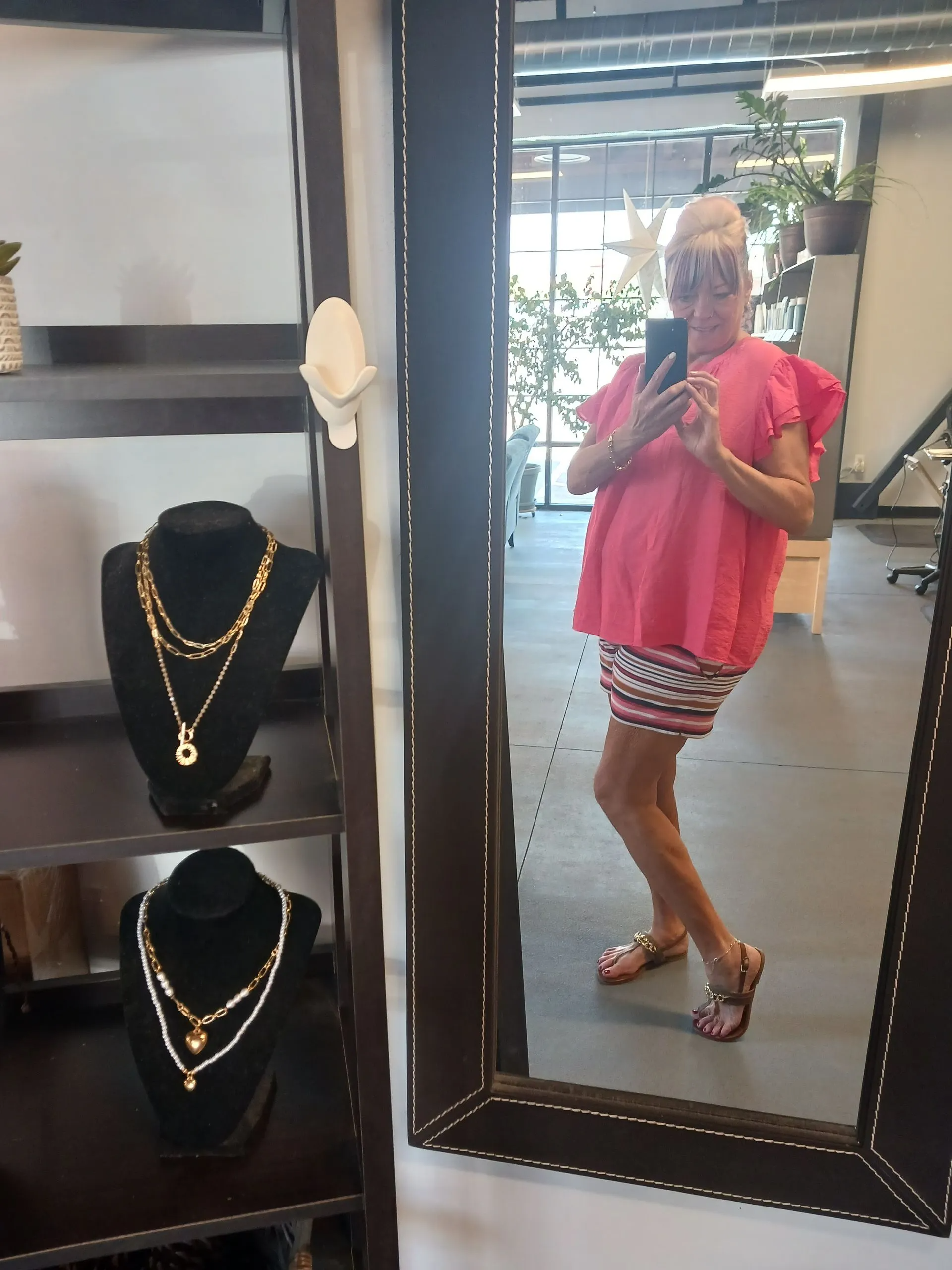 A woman takes a mirror selfie inside a modern boutique or salon while wearing a bright pink blouse, striped skirt, and sandals. Jewelry displays and indoor plants surround the stylish space, creating a fashionable, relaxed atmosphere that highlights personal style, confidence, and casual elegance.