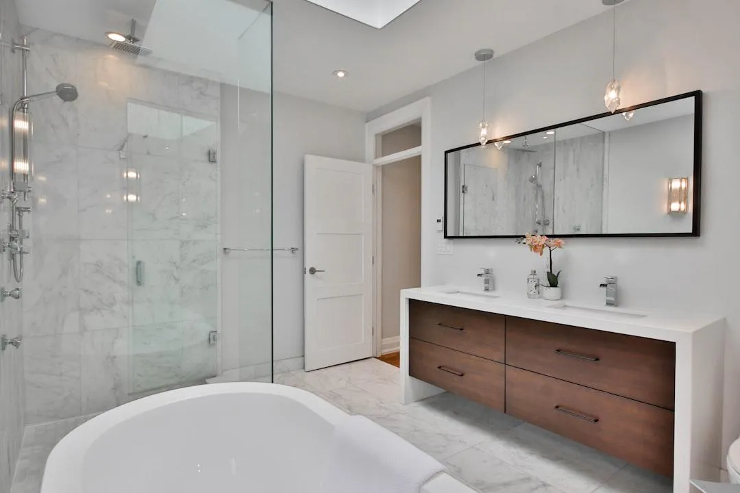 A luxurious modern bathroom with a freestanding bathtub, a glass-enclosed shower, and a double vanity with a large mirror. The bathroom features marble tile flooring and walls, and warm wood cabinetry for the vanity. Sleek chrome fixtures and contemporary pendant lights complete the sophisticated look.