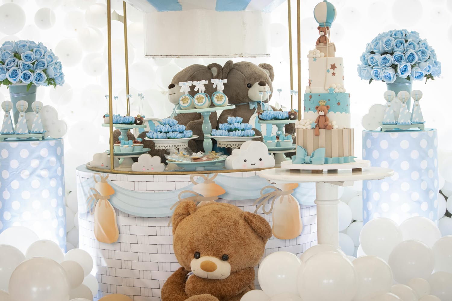 A teddy bear themed celebration table features a tiered cake, cupcakes, and other sweets decorated in blue and white. Two large teddy bears are positioned behind the cake, and a plush teddy bear sits in front of the display.