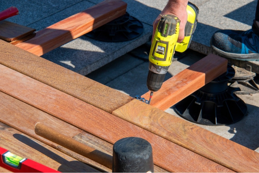 This close-up image captures the process of building a quality outdoor space, showing a worker using a bright yellow power drill to secure rich, reddish-brown wood planks for a new deck. The focus on the detailed installation work&mdash;including visible supports and a level&mdash;underscores precision, craftsmanship, and attention to detail in all phases of construction. This photo represents the dedication of skilled workers creating lasting, beautiful, and stable outdoor solutions for our clients.
