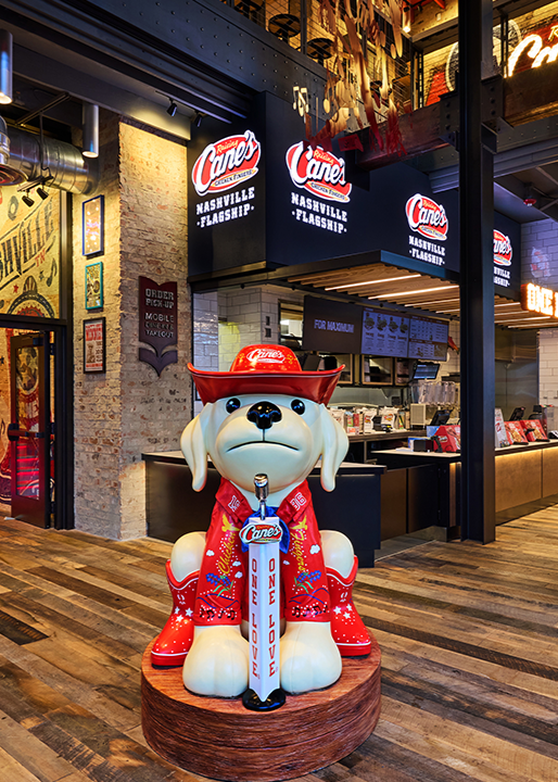 A large statue of the Raising Cane's mascot dressed in a red cowboy hat and a western shirt embroidered with "Chicken Fingers," sitting behind a microphone stand labeled "ONE LOVE" in front of the ordering counter.