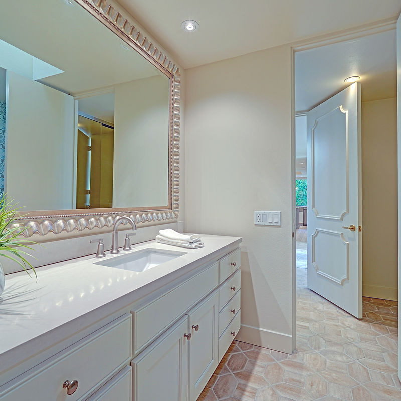 A well-lit modern bathroom features a double vanity with a large ornate mirror, a white porcelain sink, and chrome fixtures. A white towel is neatly folded on the counter. The floor is tiled in a hexagonal pattern with a wood-like finish. An open door reveals a glimpse of another room, possibly a bedroom or hallway, with more interior doors and windows.
