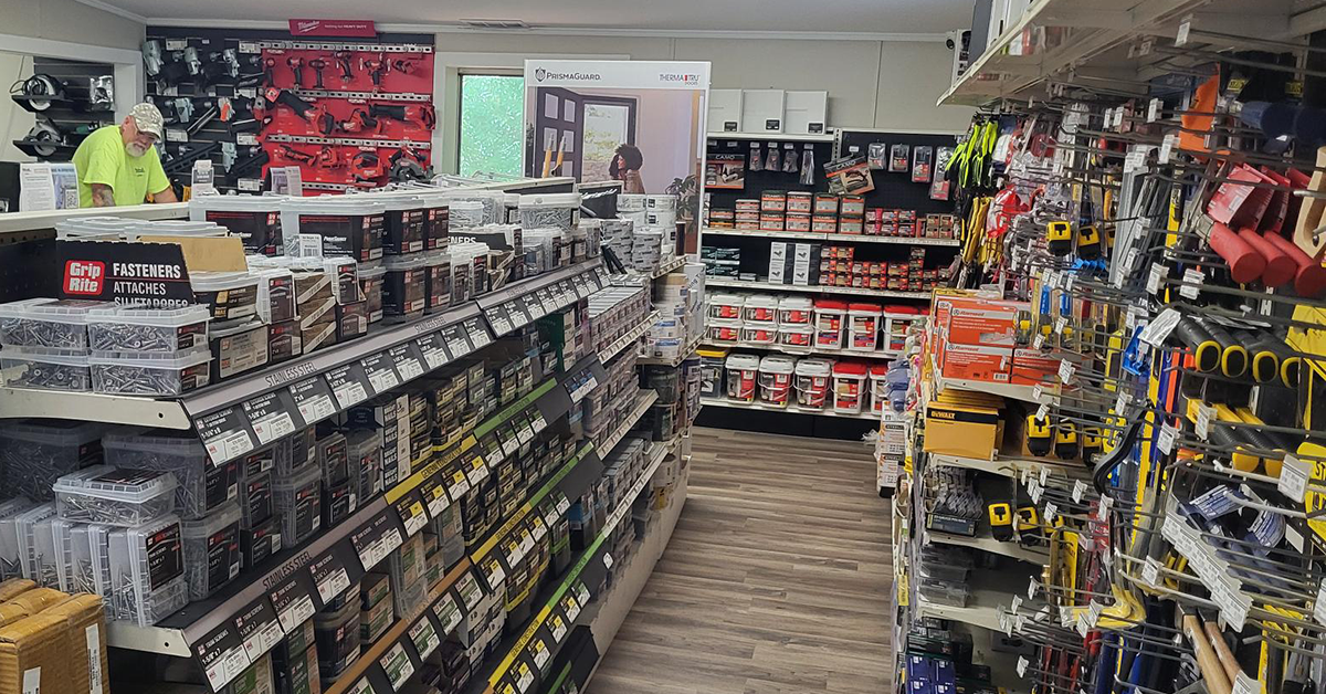 Interior showroom aisle stocked with Grip-Rite fasteners, stainless steel hardware, and roofing supplies on one side, and DeWalt tape measures, hammers, and hand tools on the other, with wall-mounted power tools and a door display visible in the background.