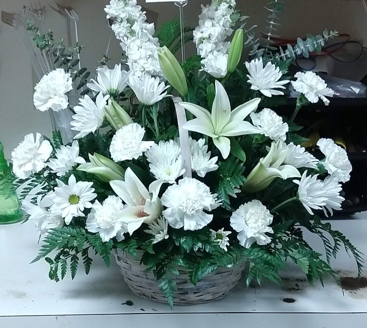 A white basket filled with all white blooms of Asiatic lilies, daisy mums, large carnation blooms, and fern foliage.