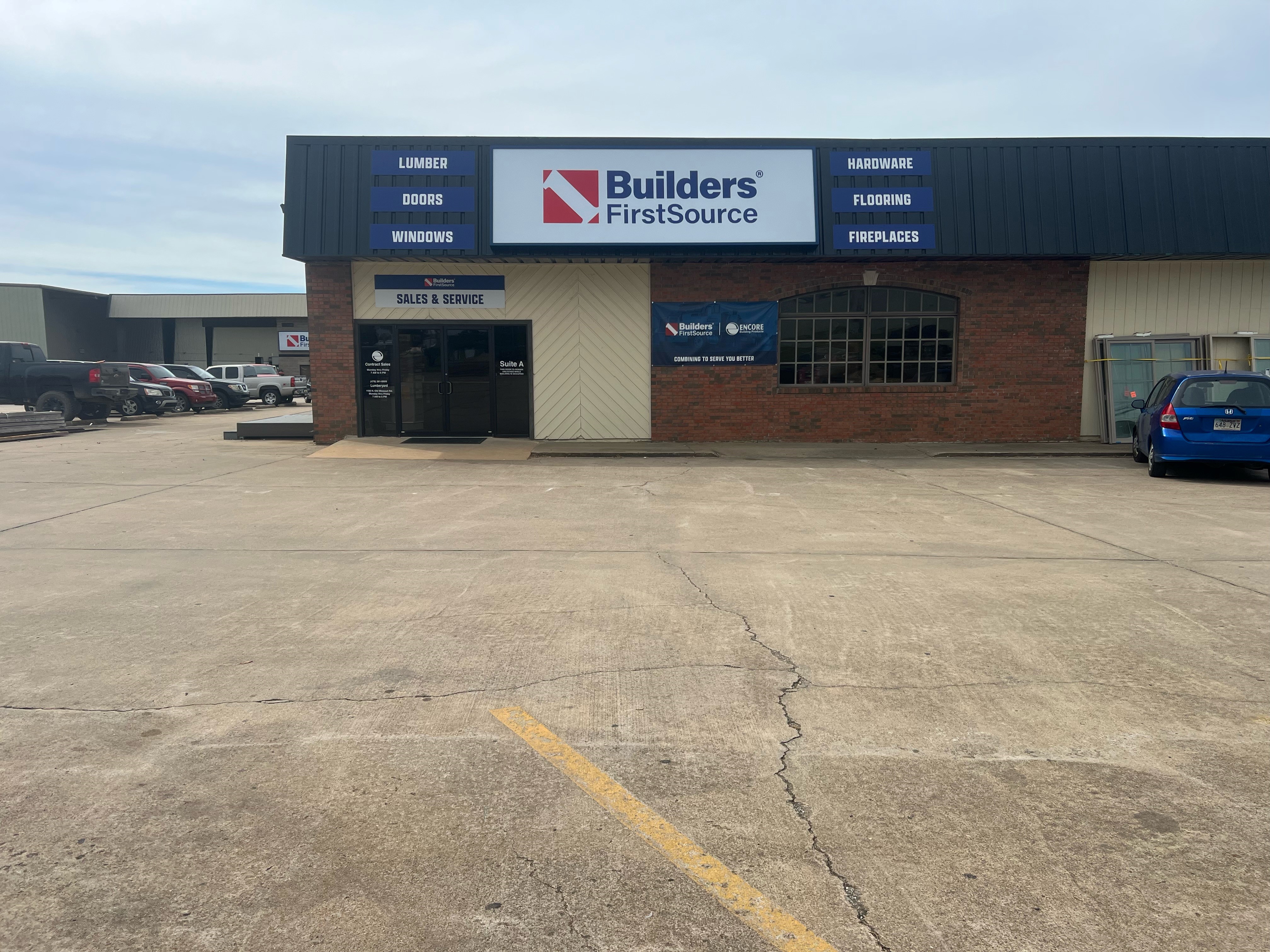 The front of a Builders FirstSource building featuring a large sign with the company logo and a list of services, including lumber, doors, windows, hardware, flooring, and fireplaces. The building has a mix of brick and light-colored siding, with a glass door marked "Sales & Service" and an adjacent window. A few vehicles are parked to the right of the building, and the surrounding area is a large, paved parking lot under a cloudy sky.