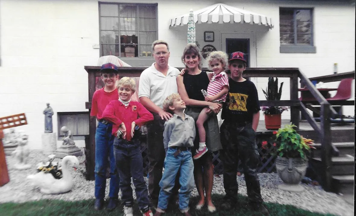 A family of seven poses for a photo in front of their house. The family includes a man, a woman, and five children. They are all smiling and appear to be happy. The house is a white two-story home with a deck and a porch. There are several decorative statues in the yard.