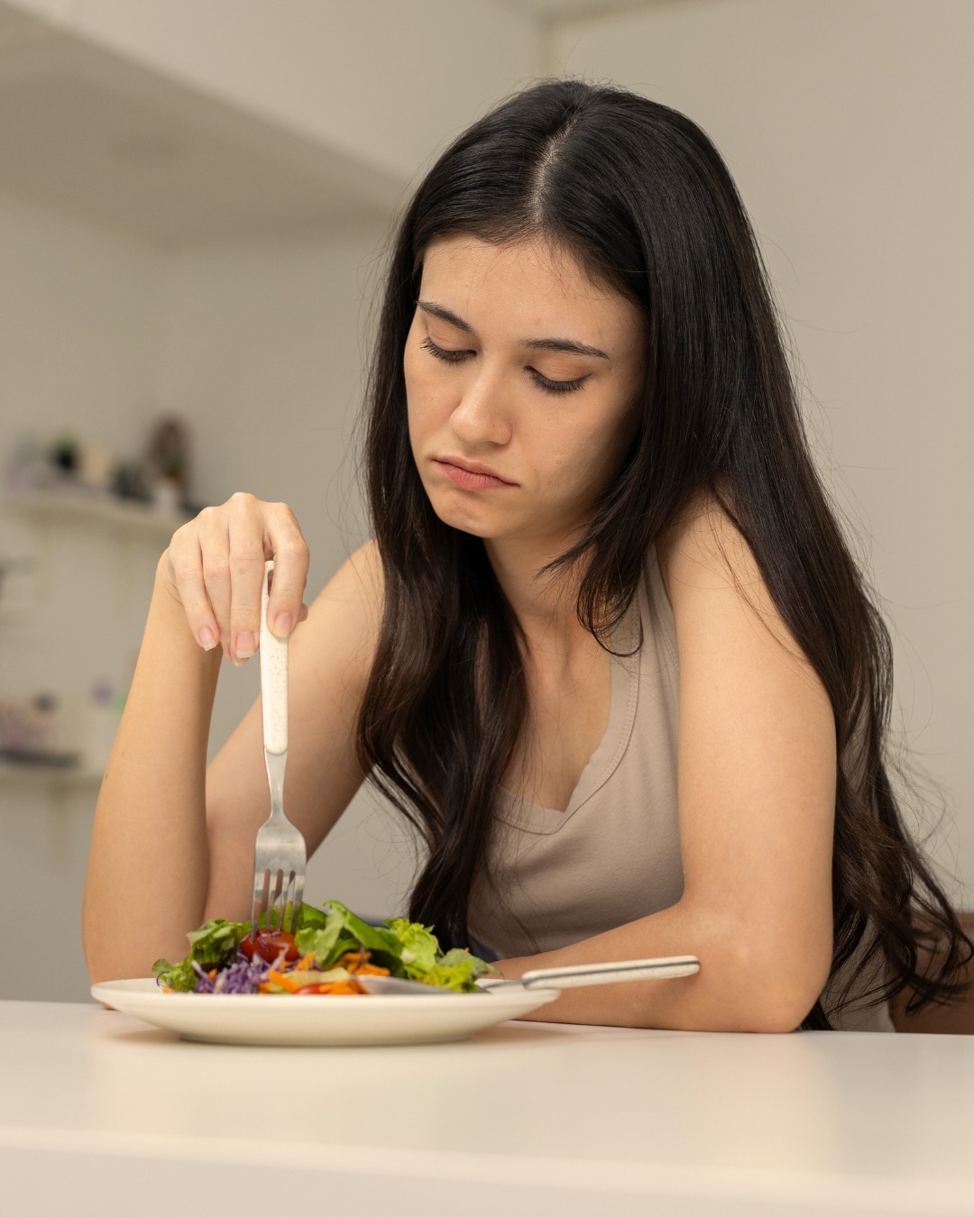 Young woman looking down at a plate of food with a serious or reluctant expression, symbolizing challenges with eating behaviors.