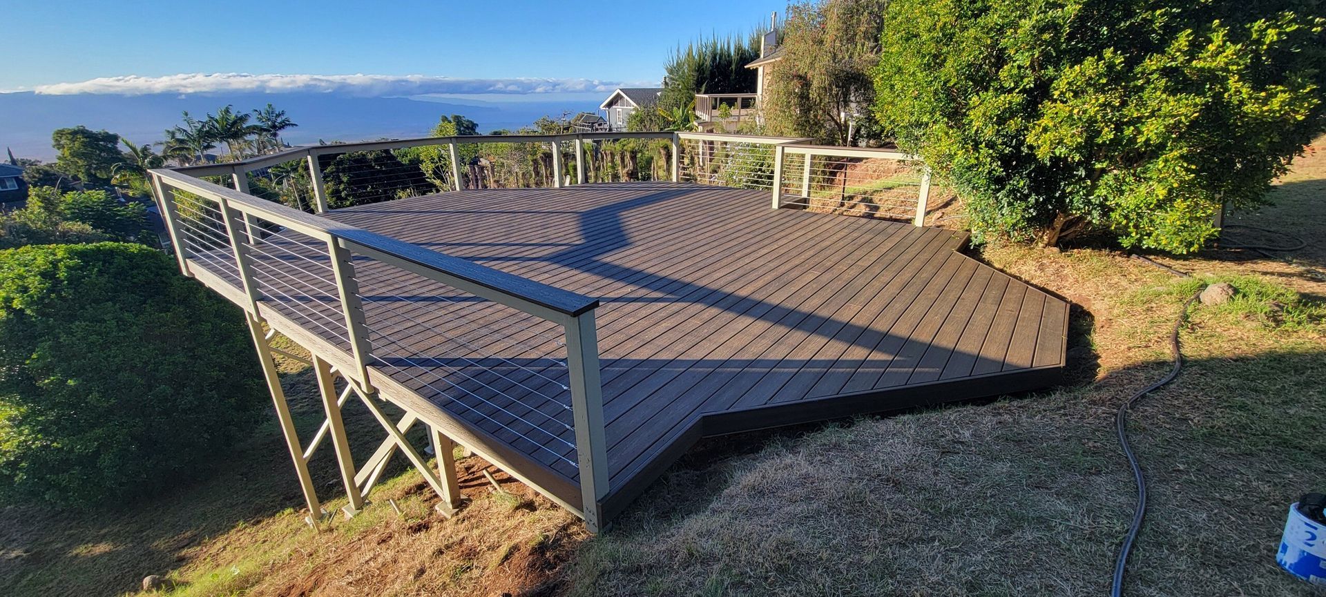 A large, elevated deck with dark composite decking overlooking a lush, mountainous view under a sunny sky. The railing features beige posts and stainless steel cable infill.