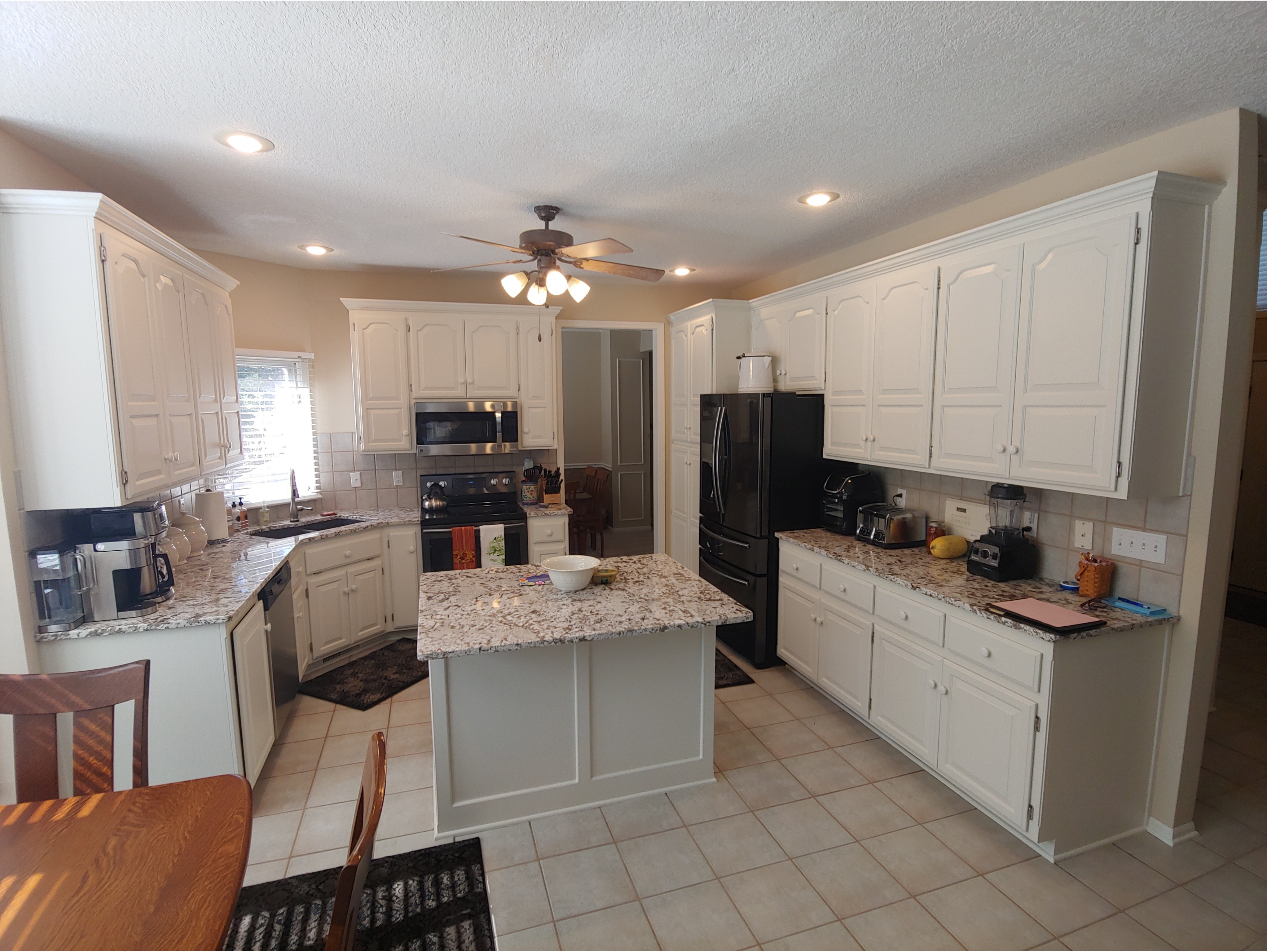 A spacious kitchen featuring freshly painted white cabinets with a smooth, updated finish, paired with granite countertops and a central island for added functionality. Perfect for showcasing kitchen cabinet painting and refinishing services that provide a modern, refreshed look while enhancing style and durability.