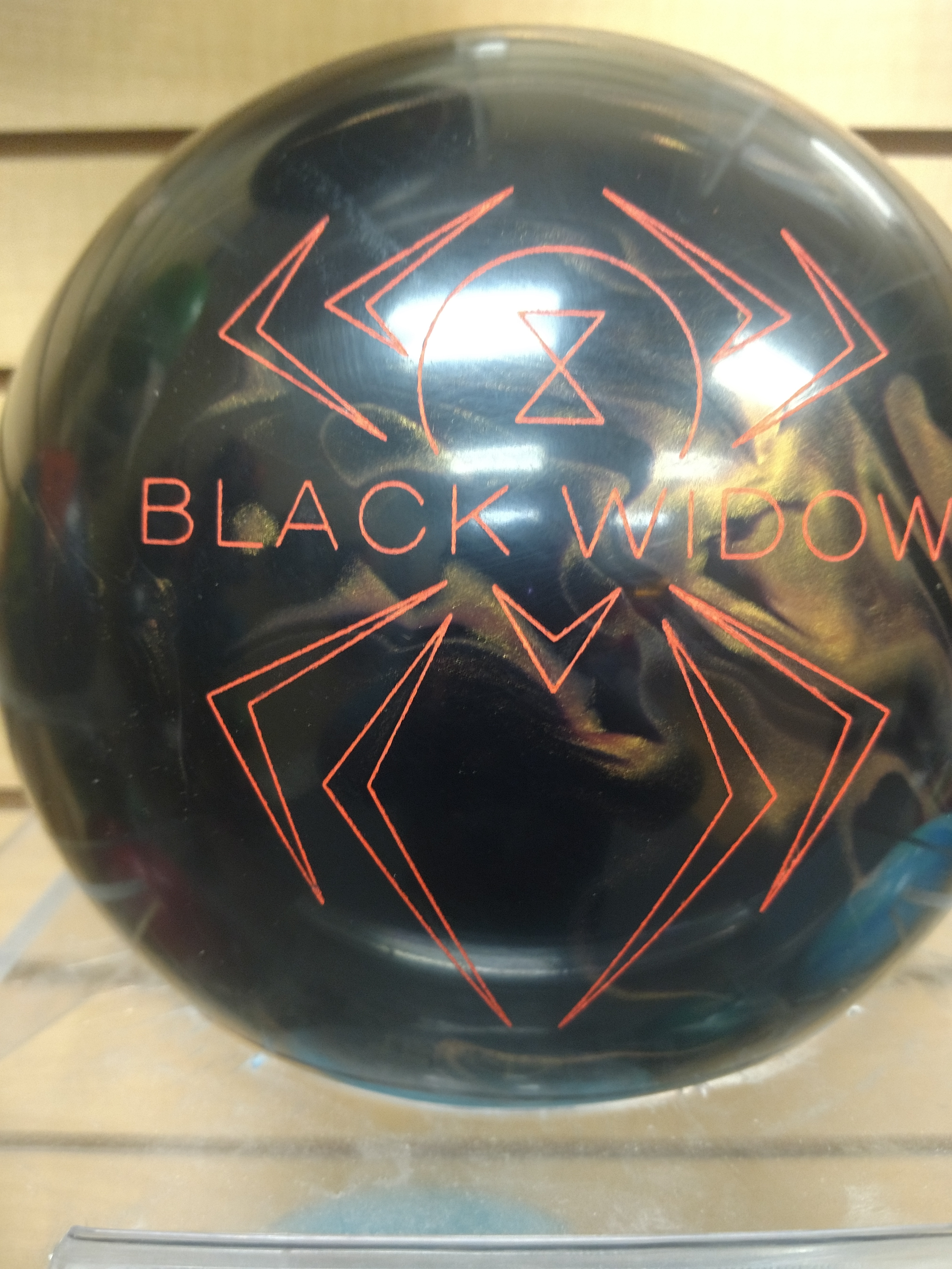 A close-up shot of a black bowling ball with bright orange graphics. The text "BLACK WIDOW" is prominently displayed in orange lettering. Above and below the text are stylized orange lines that form a spiderweb-like pattern, resembling a spider. The bowling ball has a glossy finish, reflecting light and the surrounding environment.
