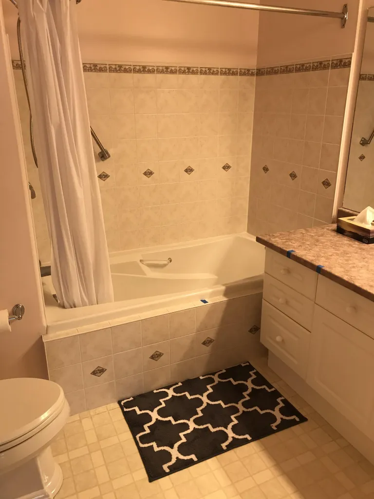 A clean bathroom with a bathtub, shower, toilet, and vanity. The walls are tiled in a cream color with decorative diamond accents. A white shower curtain hangs from a metal rod. A dark gray and white patterned rug is placed on the floor in front of the bathtub. The vanity has a speckled countertop and white cabinets with drawers. A roll of toilet paper is mounted on the wall next to the toilet.