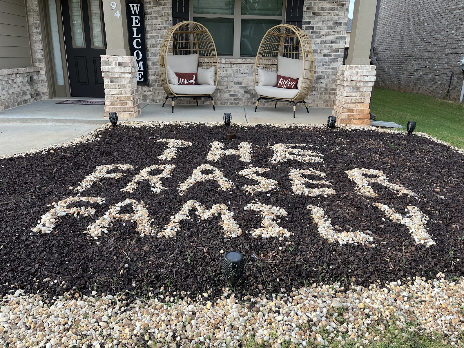 The front of a house decorated with two wicker egg chairs and a garden bed displaying the words "THE FRASER FAMILY" spelled out in stones. A "Welcome" sign is posted next to the front door. The home has a brick facade and is surrounded by a well-maintained lawn.