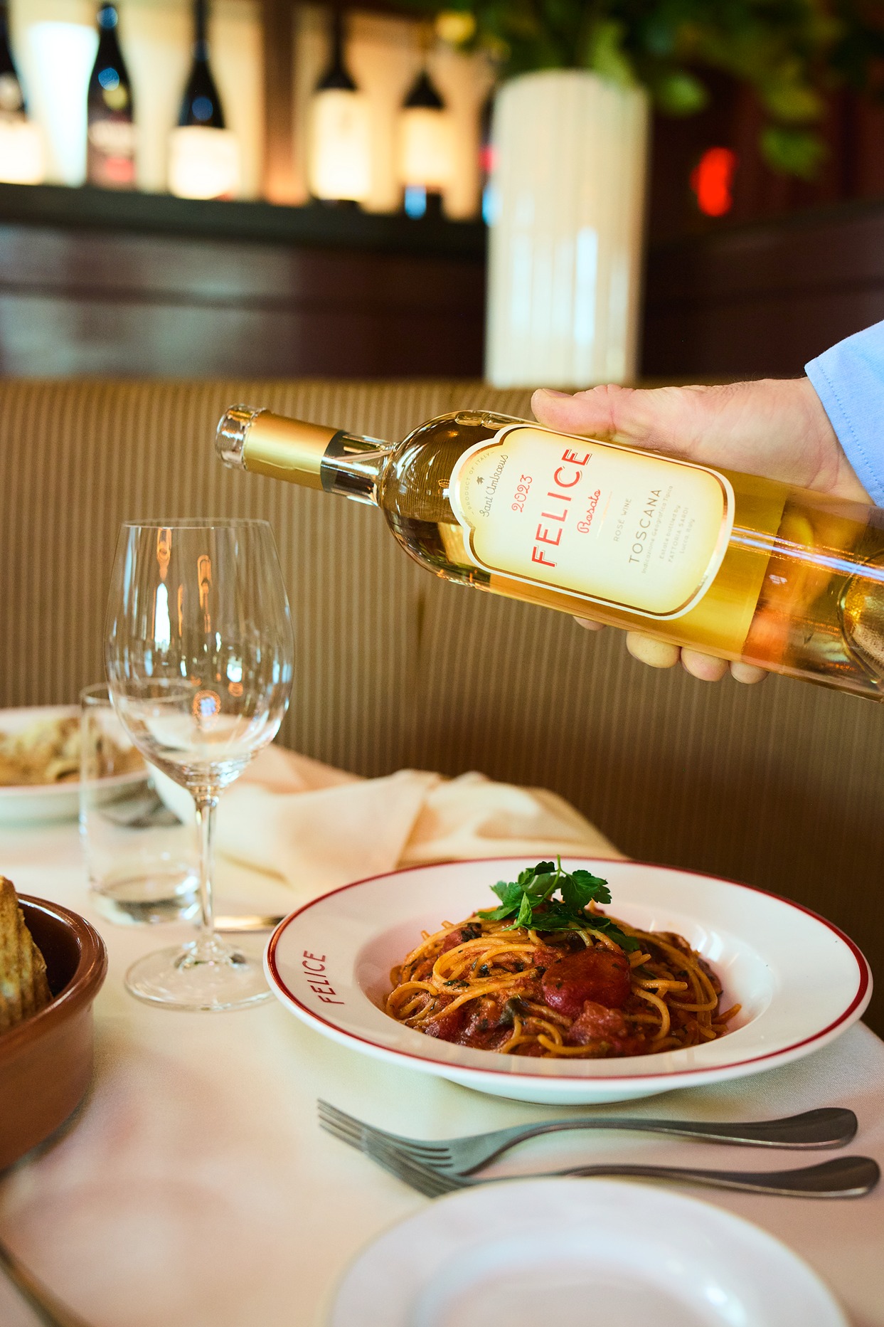 A server pours chilled wine into a glass next to a plate of spaghetti pomodoro garnished with parsley. The background shows a cozy, upscale booth.