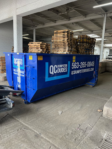 A QC Dumpster Dudes dumpster parked inside a warehouse filled with wooden pallets, ideal for industrial or commercial debris removal.