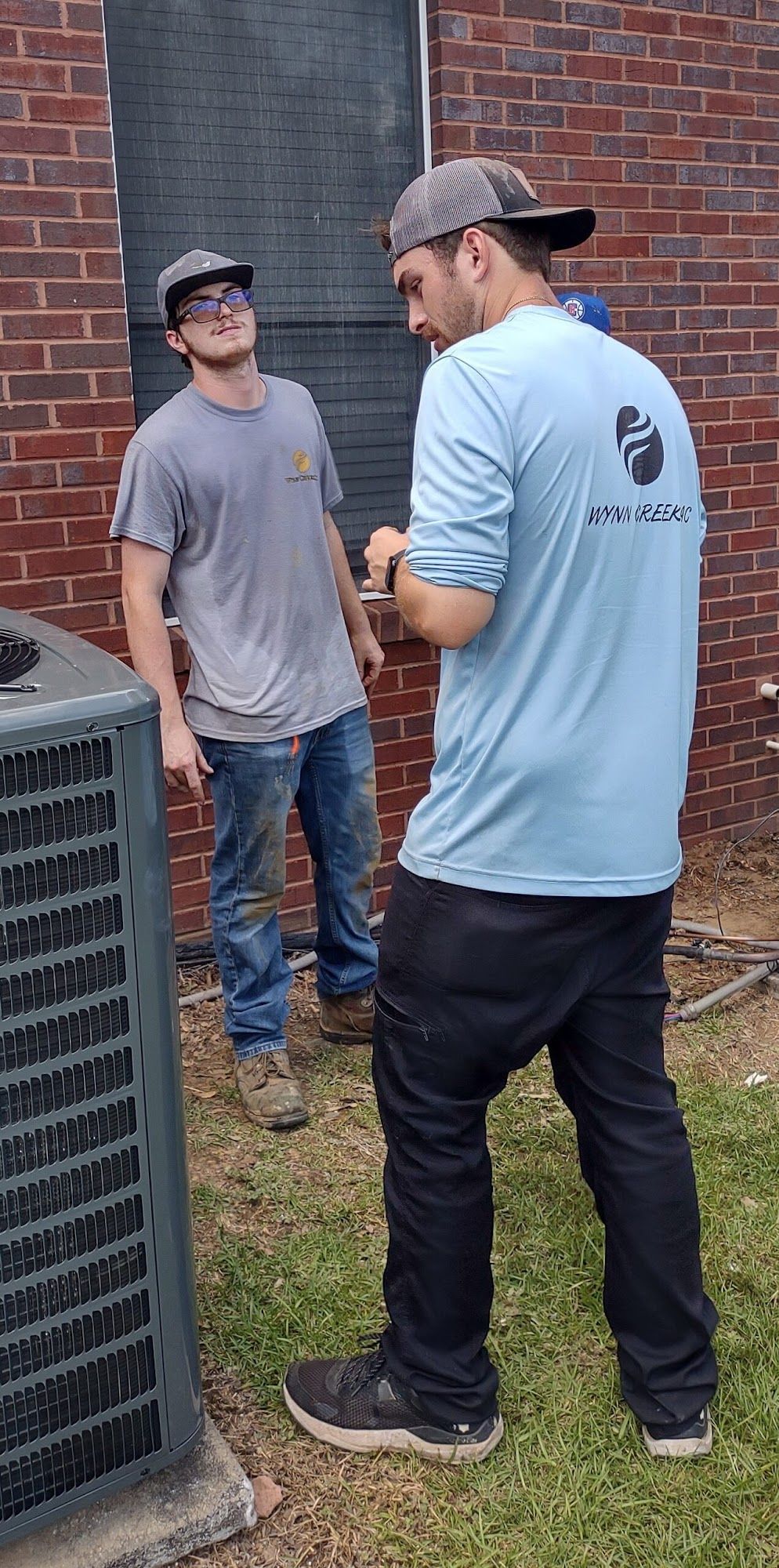 Two HVAC technicians discussing maintenance near outdoor air conditioning unit beside brick wall, reviewing system performance and service needs, highlighting teamwork, inspection process, and residential cooling system support.