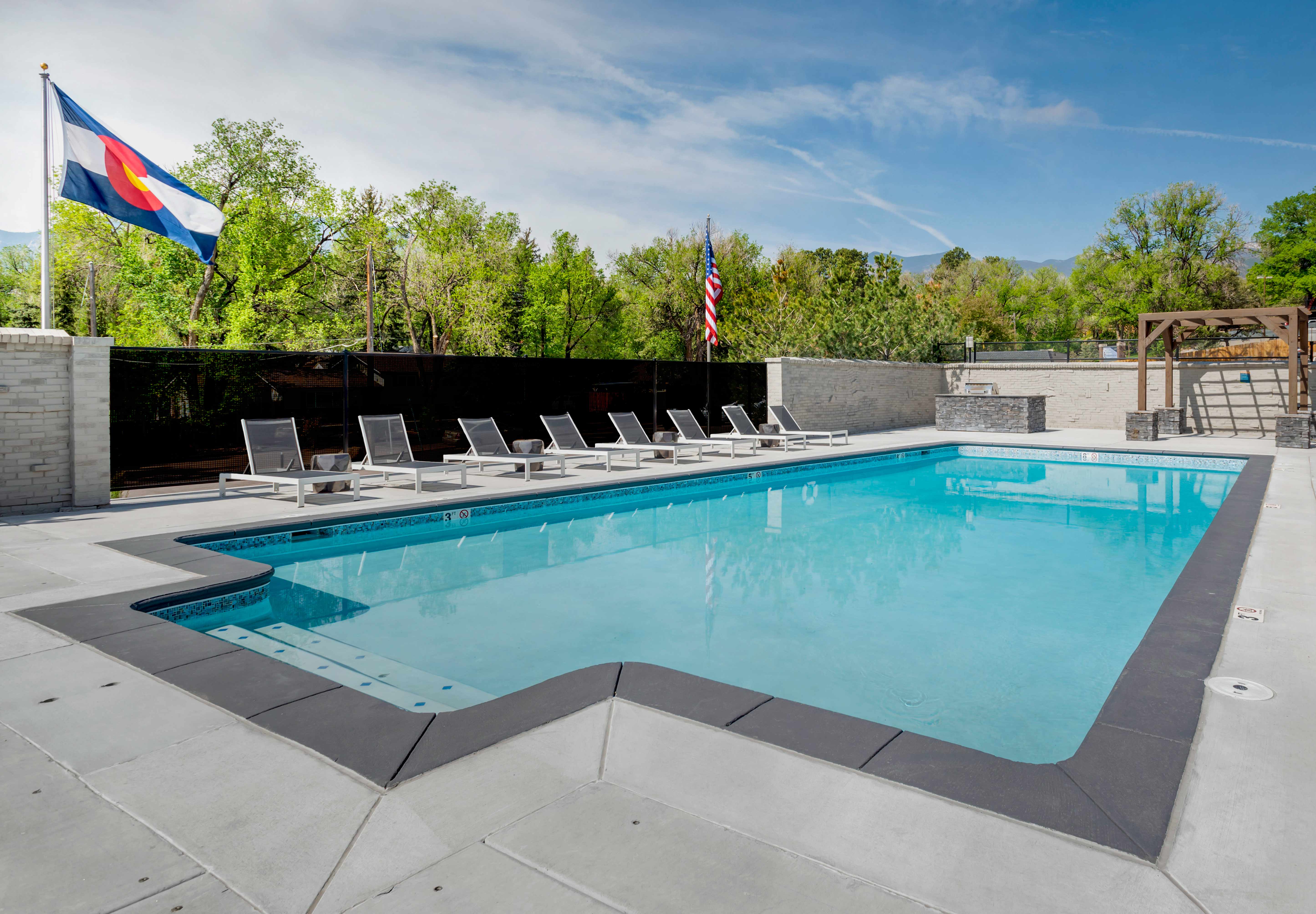 Rectangular pool with lounge seating, gas grill, pergola, and Colorado flag.