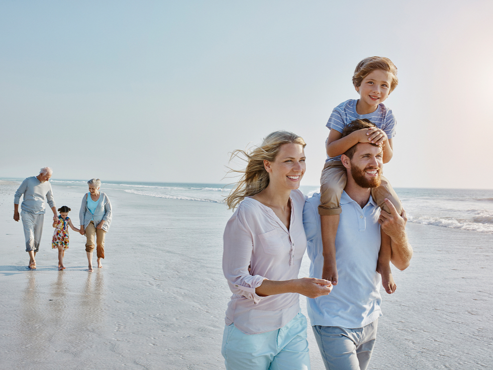 A family enjoys a sunny day on the beach. A man carries a smiling child on his shoulders, walking alongside a woman. An elderly couple and child stroll behind them.
