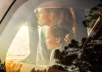 A mother and daughter looking at a calm lake as seen through a reflective car windshield.