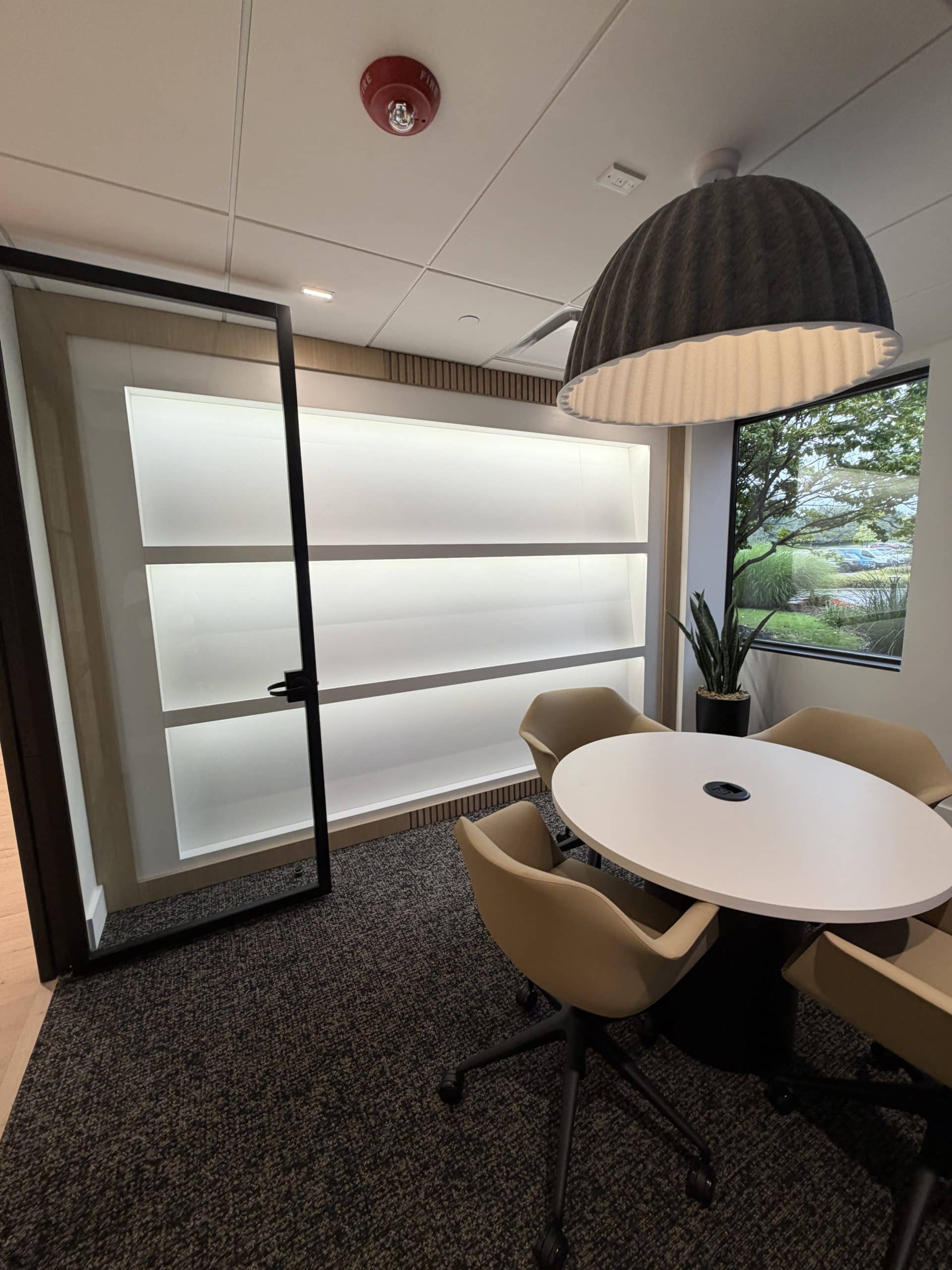 A welcoming meeting room at the Toll Brothers Michigan Division Office & Design Studio showcases the attention to interior detail within Schonsheck, Inc.’s commercial construction portfolio. Glass enclosure, illuminated wall shelving, and a distinctive pendant light give the space an open, polished appearance. A round table and upholstered seating provide a comfortable setting for small meetings and conversations, bringing practical function and visual appeal together in a professional office environment.