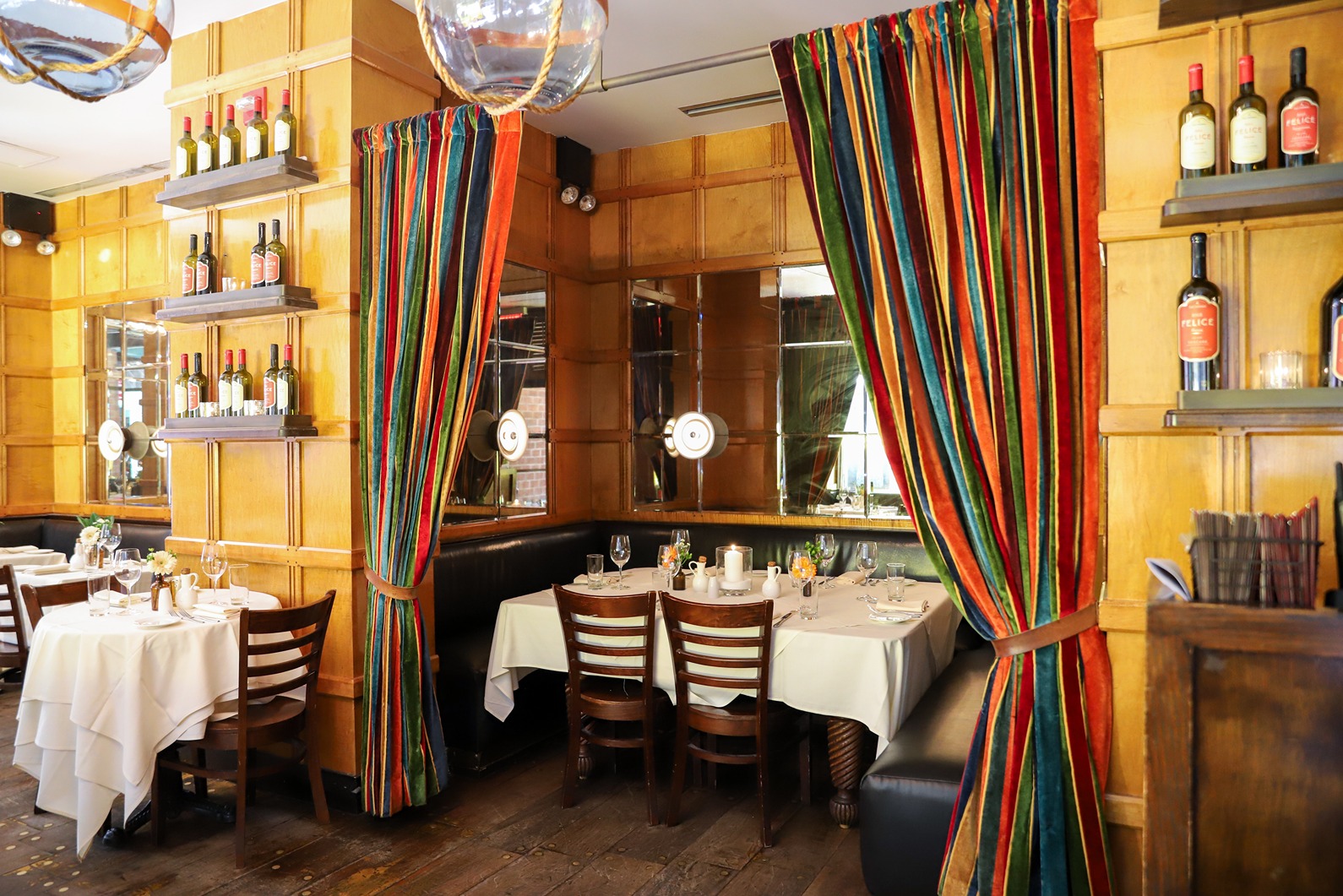 Warm wood paneling and mirrors surround intimate dining booths with white tablecloths. Vibrant, multi-colored striped velvet curtains are draped to the side, adding a rich texture to the rustic wood floors and wine bottle displays.