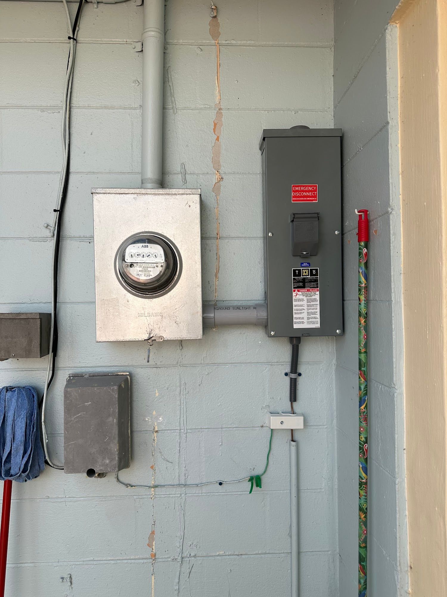 Exterior electrical setup mounted on a painted cinder block wall featuring a utility meter, emergency disconnect panel, conduit piping, and wiring connections, representing residential electrical service equipment and safety components.
