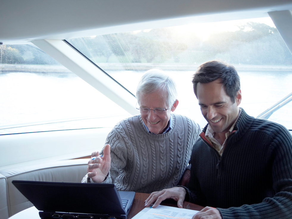 Two men smiling and engaged in using a laptop on a boat. One gestures at the screen, wearing a gray sweater, while the other wears a dark sweater. Sunlight streams through large windows, and a serene water view is visible.