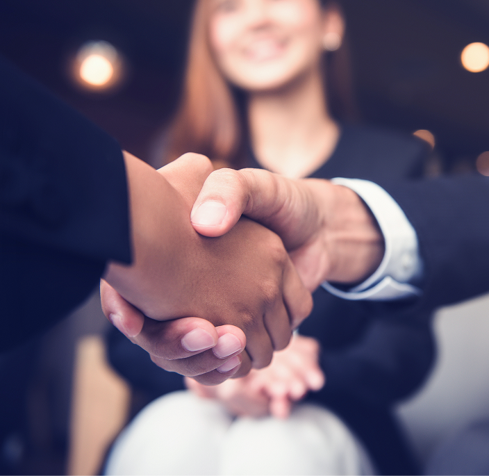 Close up of two professionals shaking hands while a third person looks on, smiling.