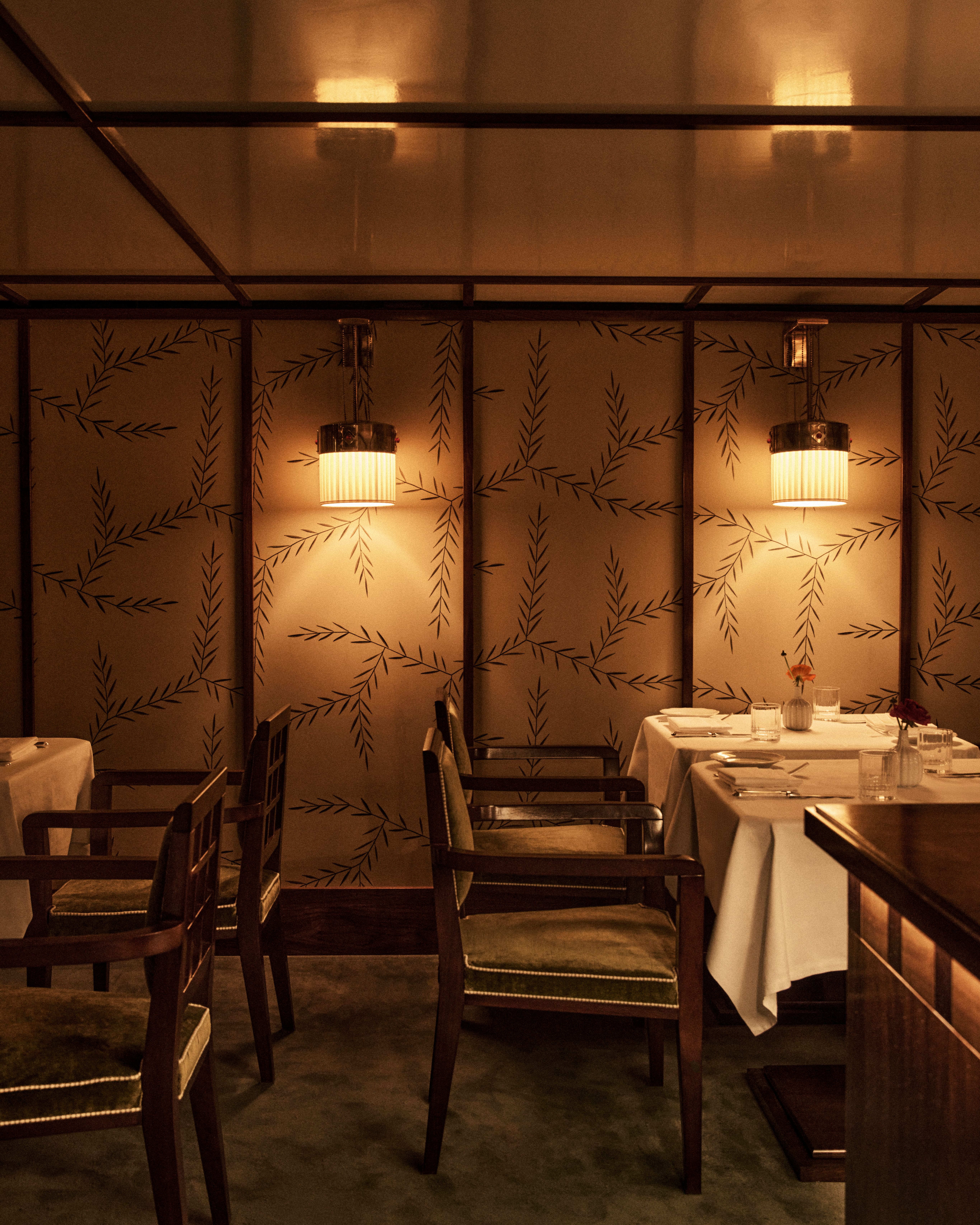 Cozy restaurant interior with small tables dressed in white linens, dark wooden chairs with green upholstery, softly glowing pendant lights, and patterned wall panels creating a warm, elegant atmosphere.