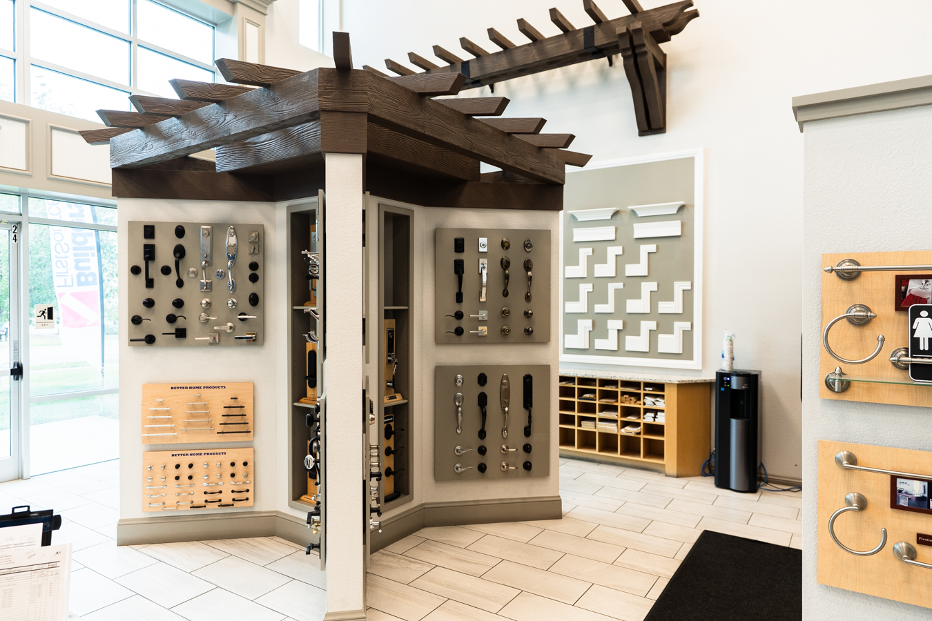 Builders FirstSource showroom with hardware displays, a wooden pergola, and molding samples on a tiled floor.