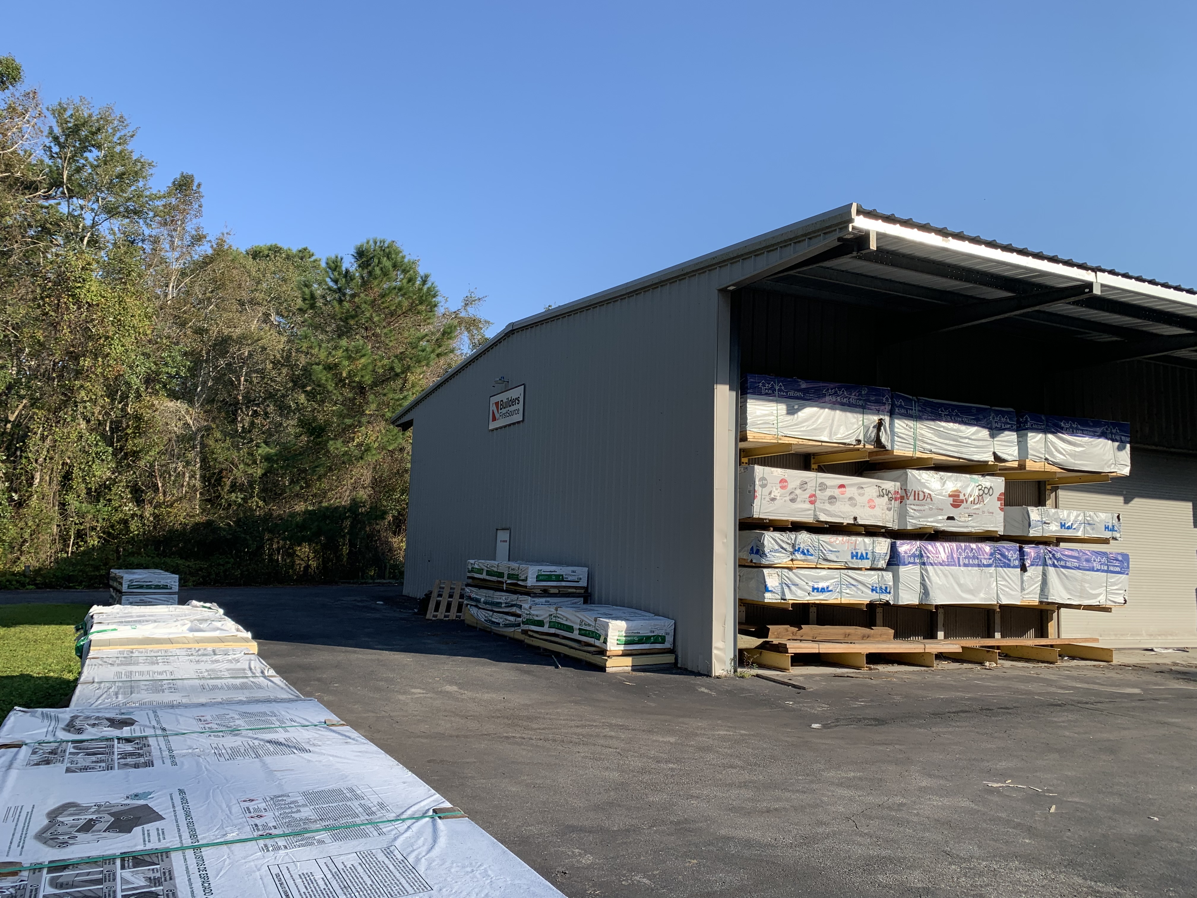 n exterior shot of a warehouse with stacks of lumber and building materials visible under a roofed area. The building is labeled "Builders FirstSource" on the side. Bundled stacks of wood and other materials are arranged along the paved ground outside the warehouse, with trees and greenery in the background. The sky is clear and blue.