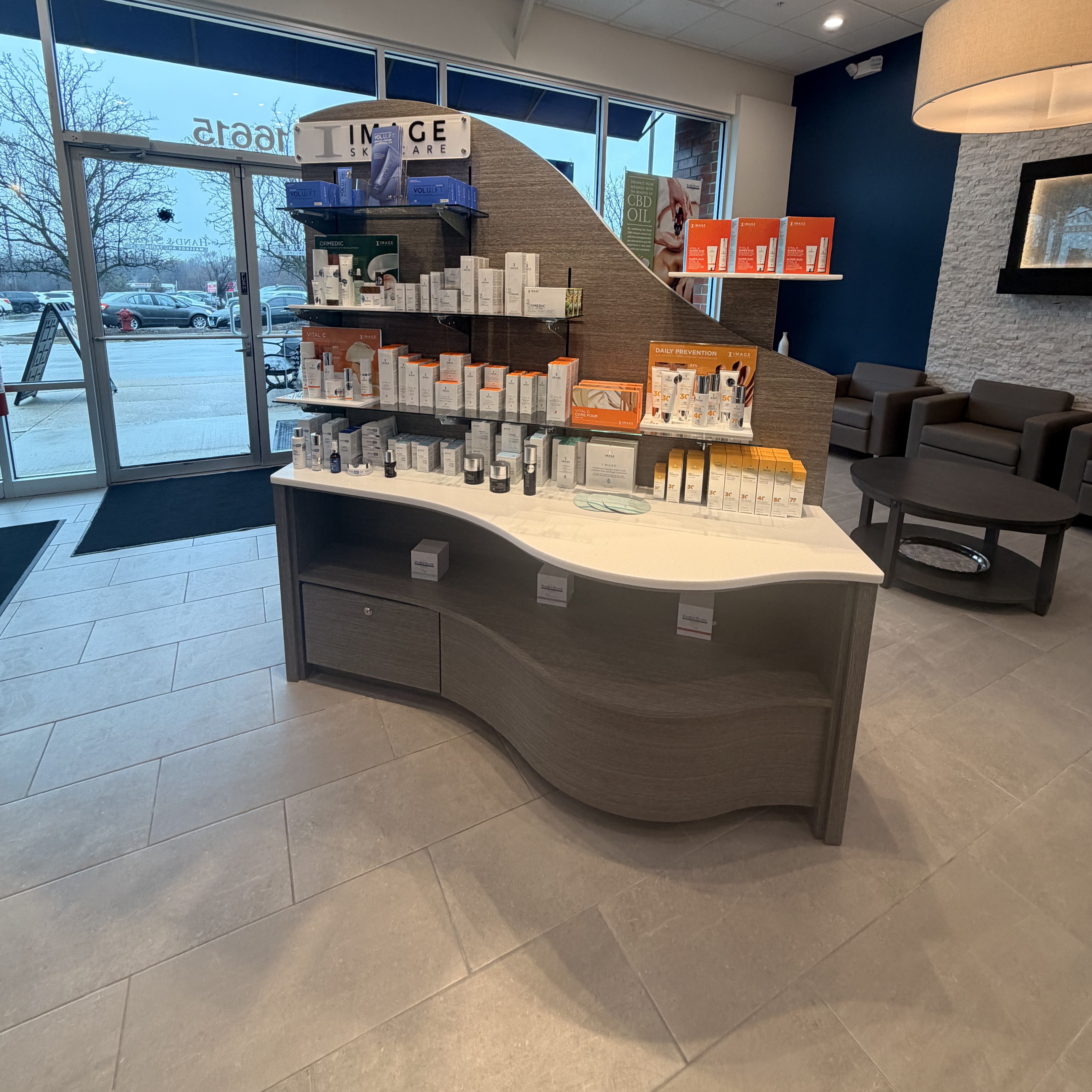 Curved reception desk in gray wood-grain finish with white countertop. Glass shelving displays skincare products in orange, white, and blue packaging. Gray upholstered seating visible. Large windows show exterior street view. White ceiling with recessed lighting and exit sign.