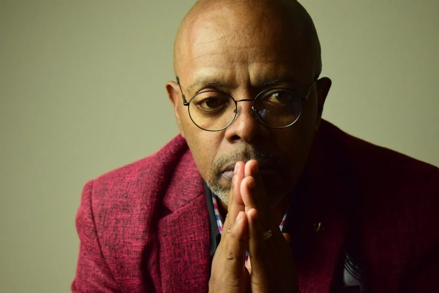 A portrait of a bald Black man wearing glasses and a deep red textured blazer, with his hands clasped together in front of his mouth. He appears to be looking directly at the camera with a contemplative expression. The background is a neutral, muted green-grey.