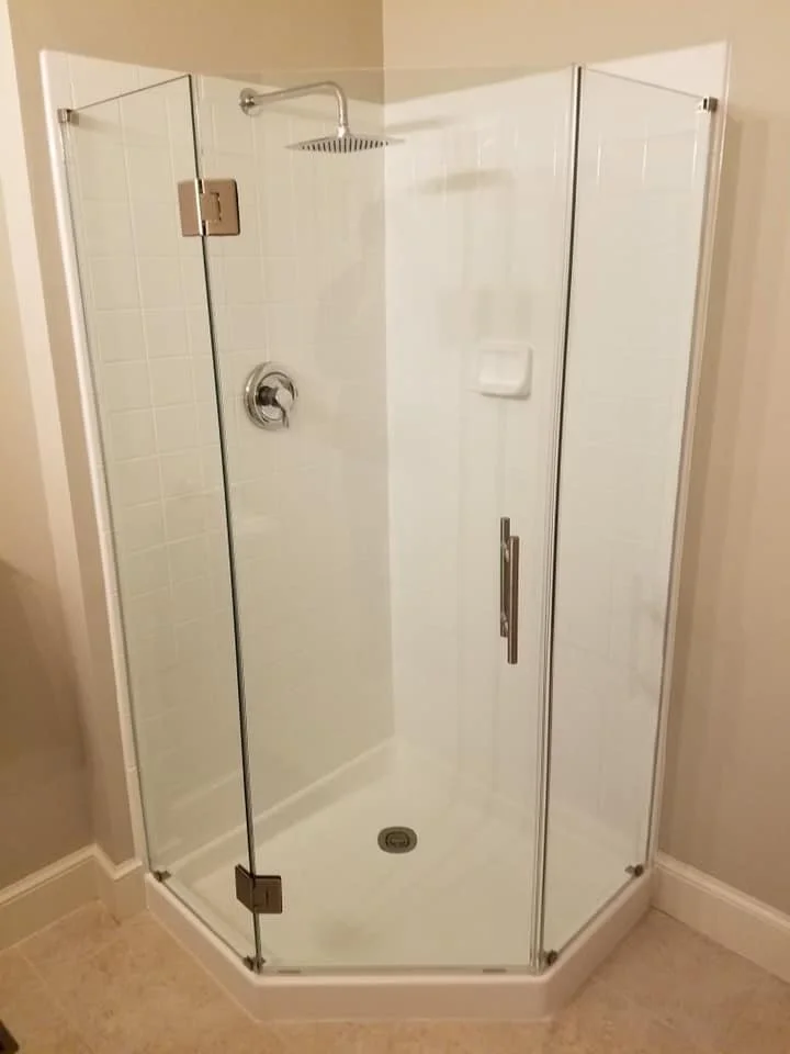 A neo-angle shower enclosure with glass doors and white tiled walls is shown. The shower features a chrome showerhead, a shower valve, a soap dish, and a drain in the base. The enclosure is set in a bathroom with beige walls and tiled flooring.