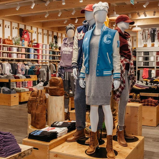 Inside a Roots Location, a mannequin dressed in a blue varsity jacket and short dress stands in front and the wall is lined with Roots Merchandise