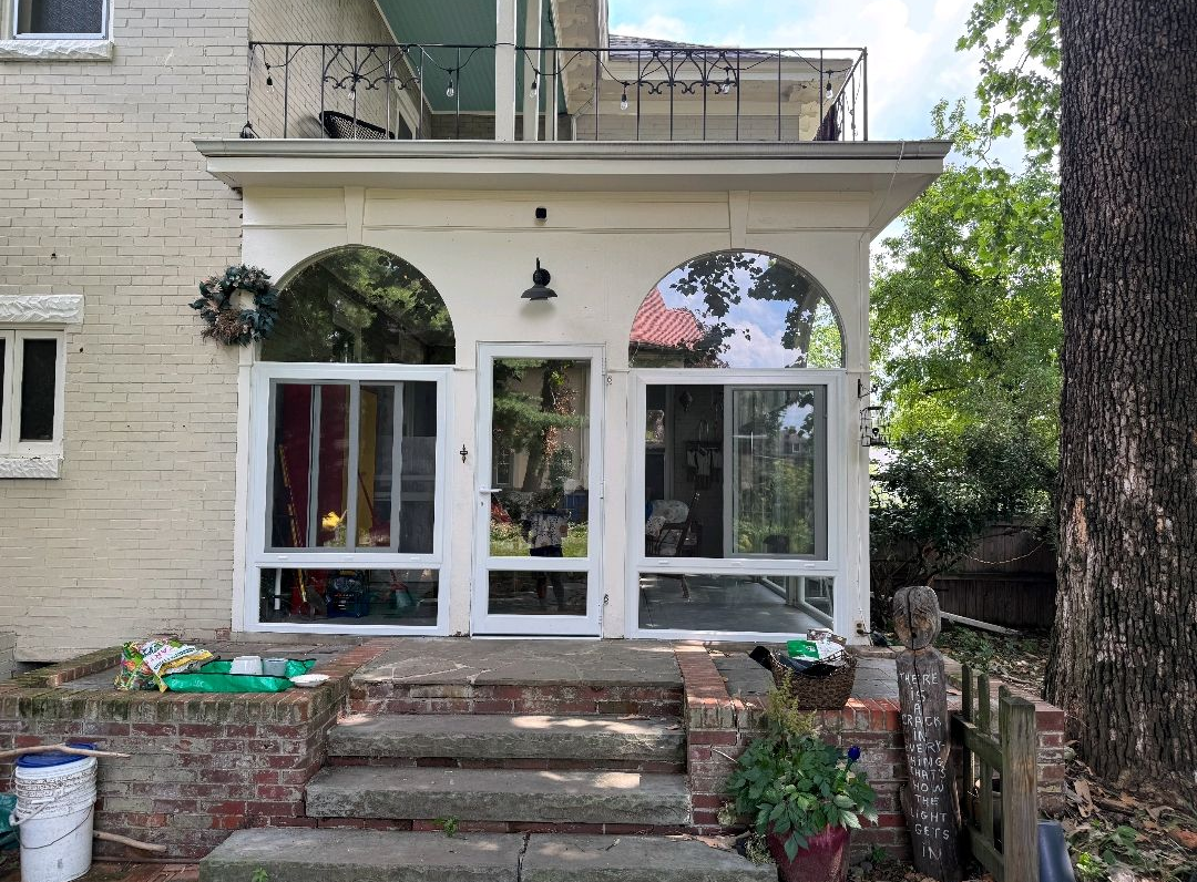 Rear view of a residential home featuring a screened-in patio with large glass panels, arched upper windows, and a central door. Brick steps lead to the entrance, with surrounding greenery and a balcony above enhancing outdoor living space.